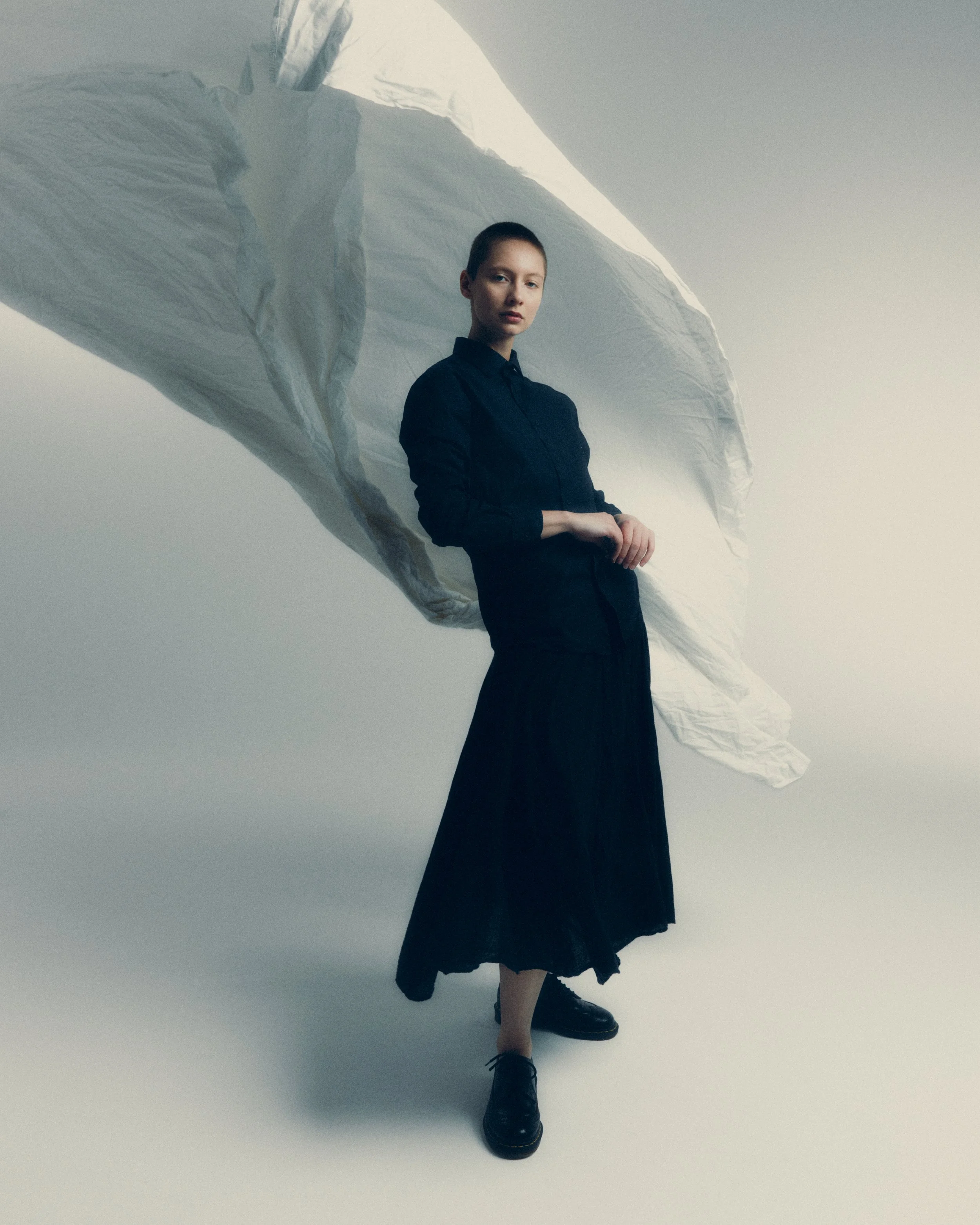 A woman standing on a crumpled white sheet, wearing a black long-sleeve shirt and a black skirt, with a large white fabric billowing above her head.