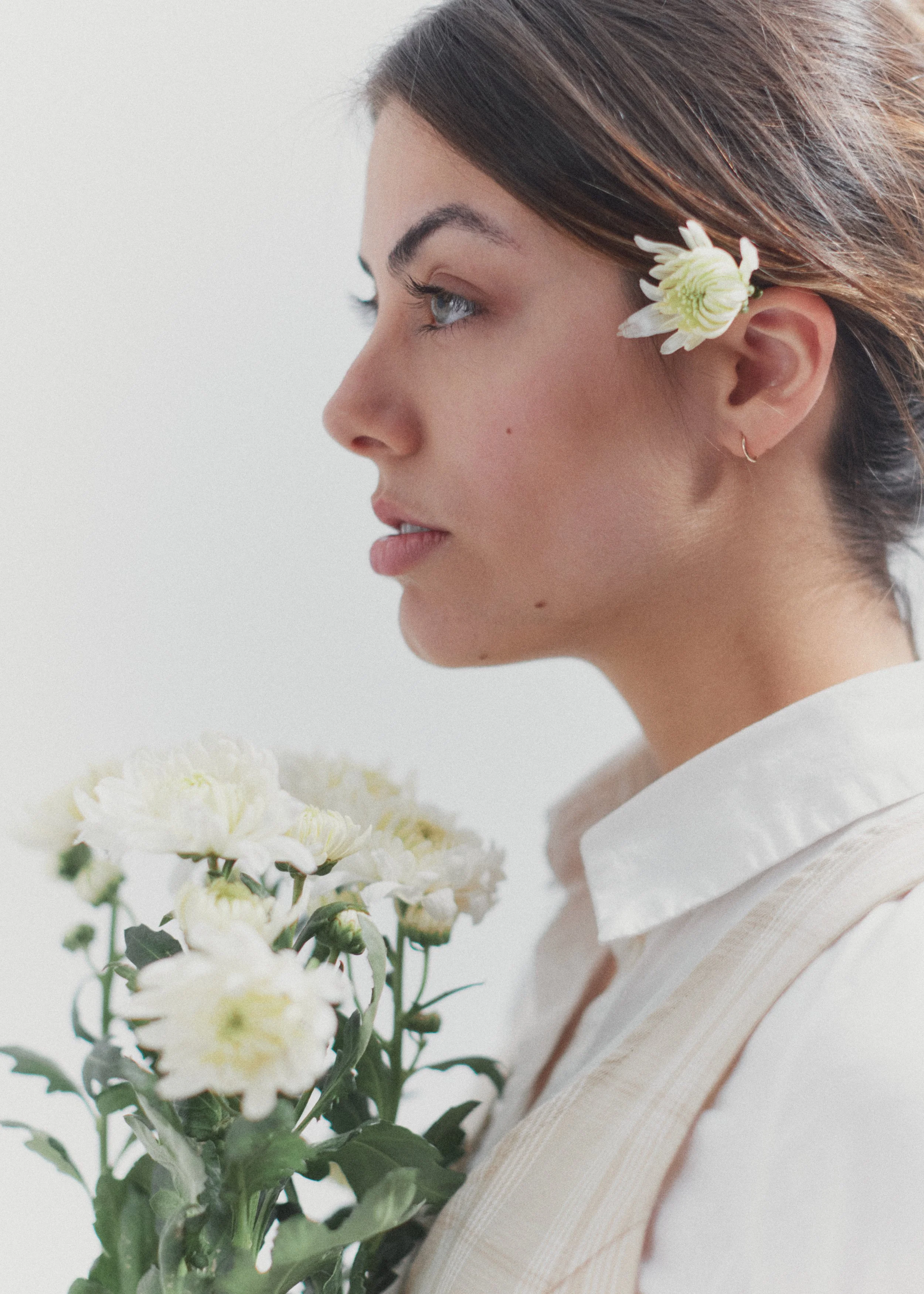 A woman with brown hair, blue eyes, and wearing a small hoop earring is holding a bouquet of white flowers. She has a white flower tucked behind her ear and is wearing a white shirt with a light beige vest. She is in profile against a plain, light ba