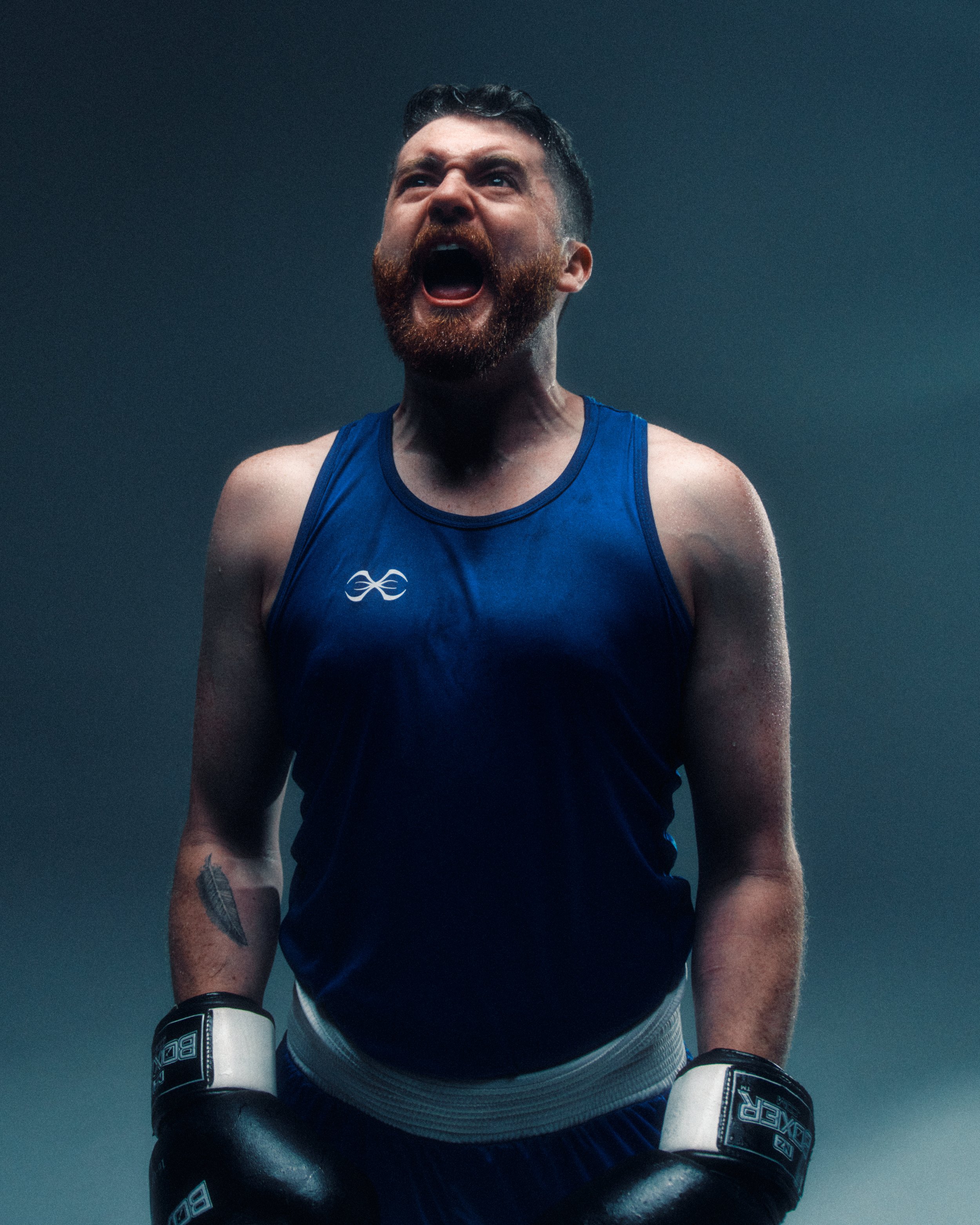 A male boxer with a beard, wearing a blue tank top and black boxing gloves, is in a fighting stance with his mouth open, against a dark, plain background.