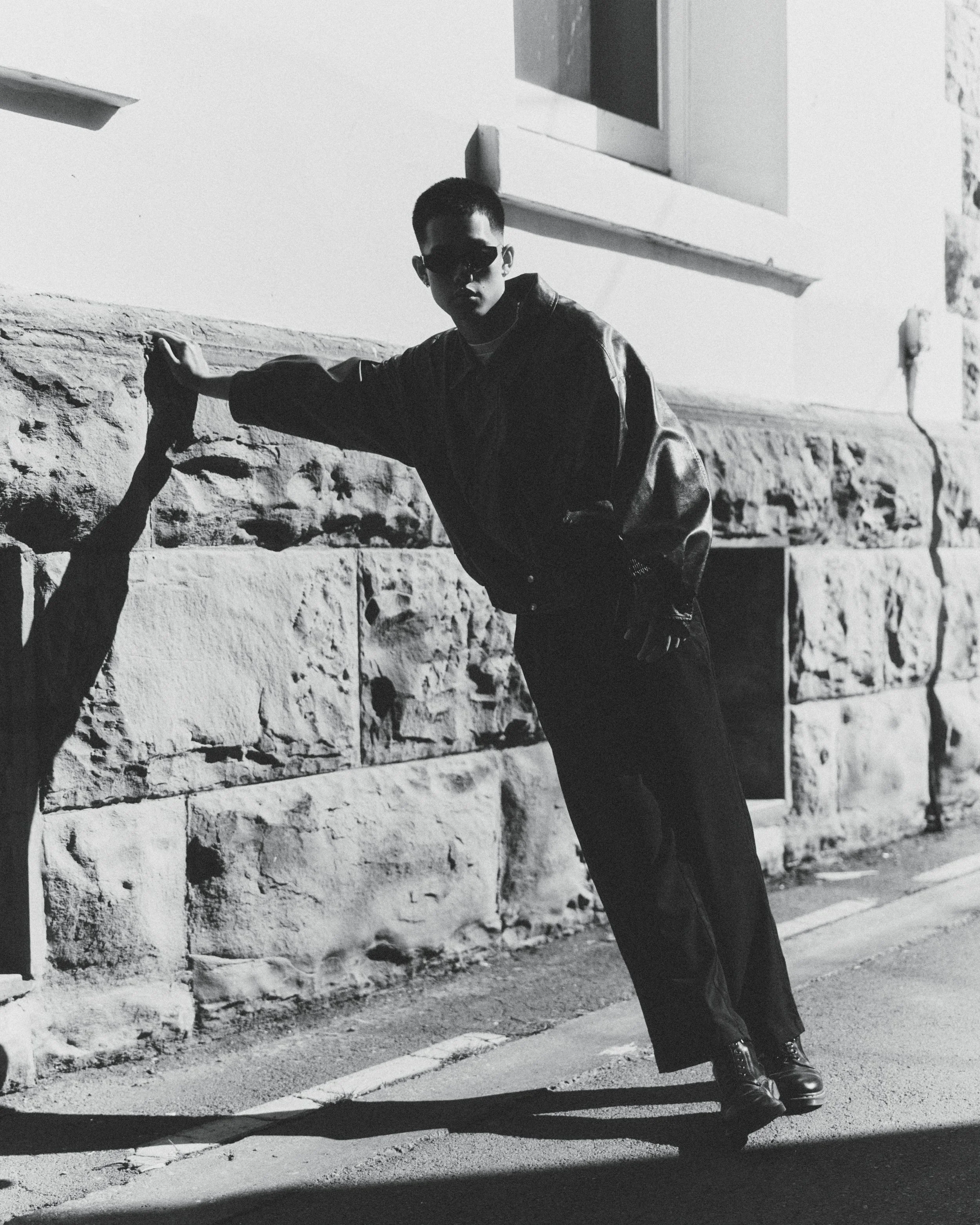 Black and white photo of a young man wearing sunglasses and a leather jacket, leaning against a stone wall with one hand, casting a shadow on the sidewalk.