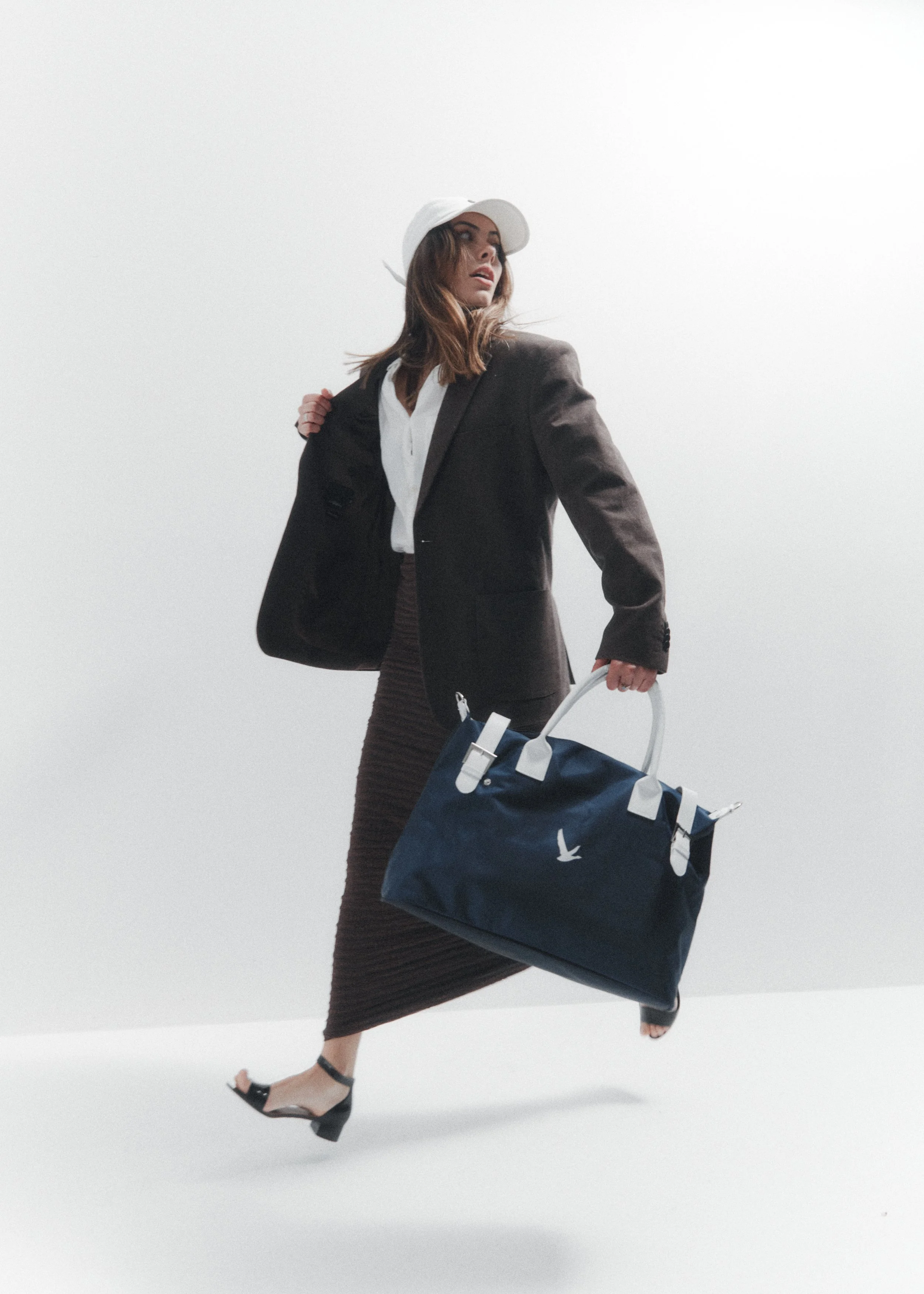 A woman dressed in a black blazer, white blouse, and a long brown skirt, wearing a white cap, holding a navy blue handbag with a white strap, and walking against a white background.