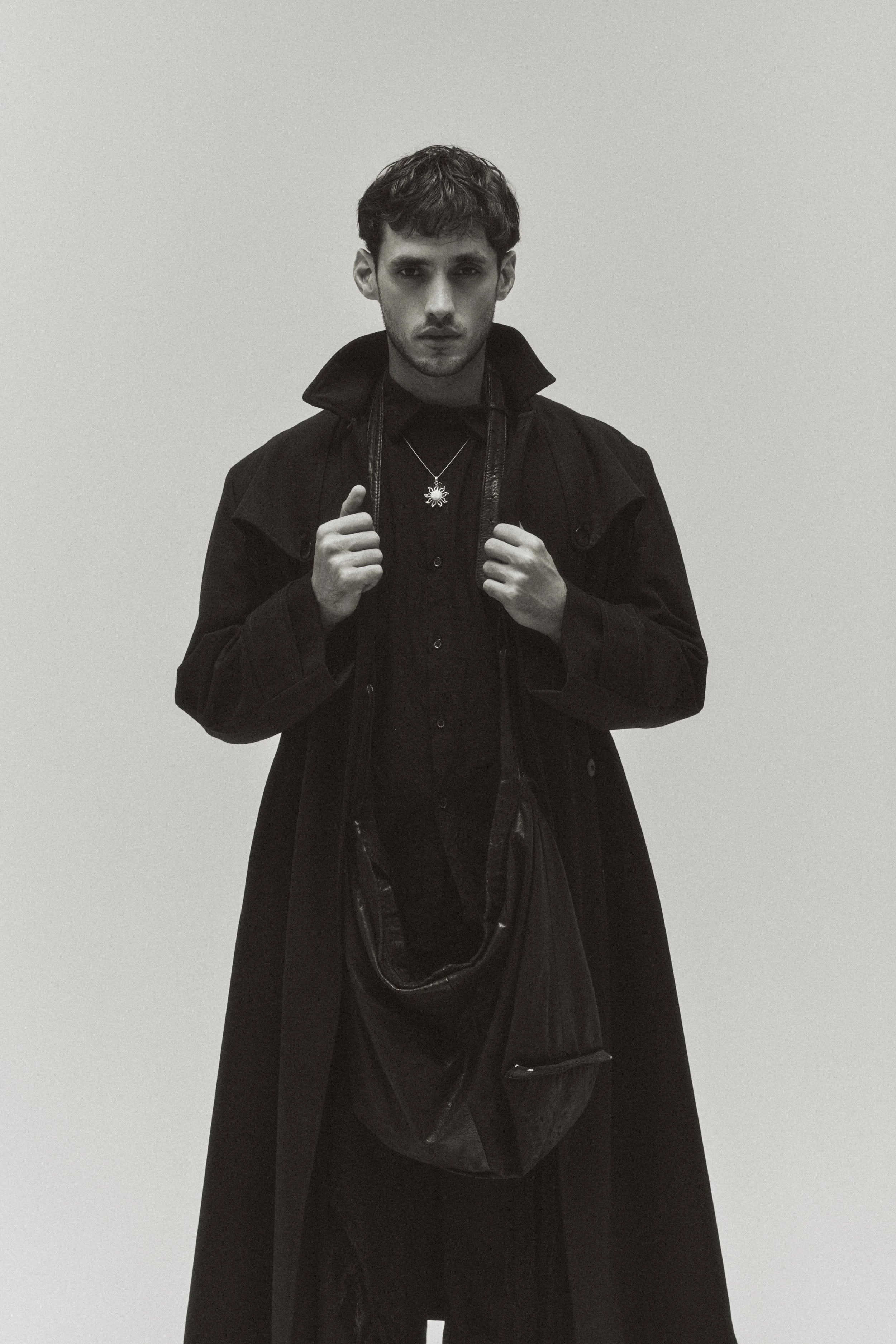 Black and white photo of a young man with wavy hair, wearing a long coat and layered black clothing, standing against a plain light background, holding straps of a bag with both hands.