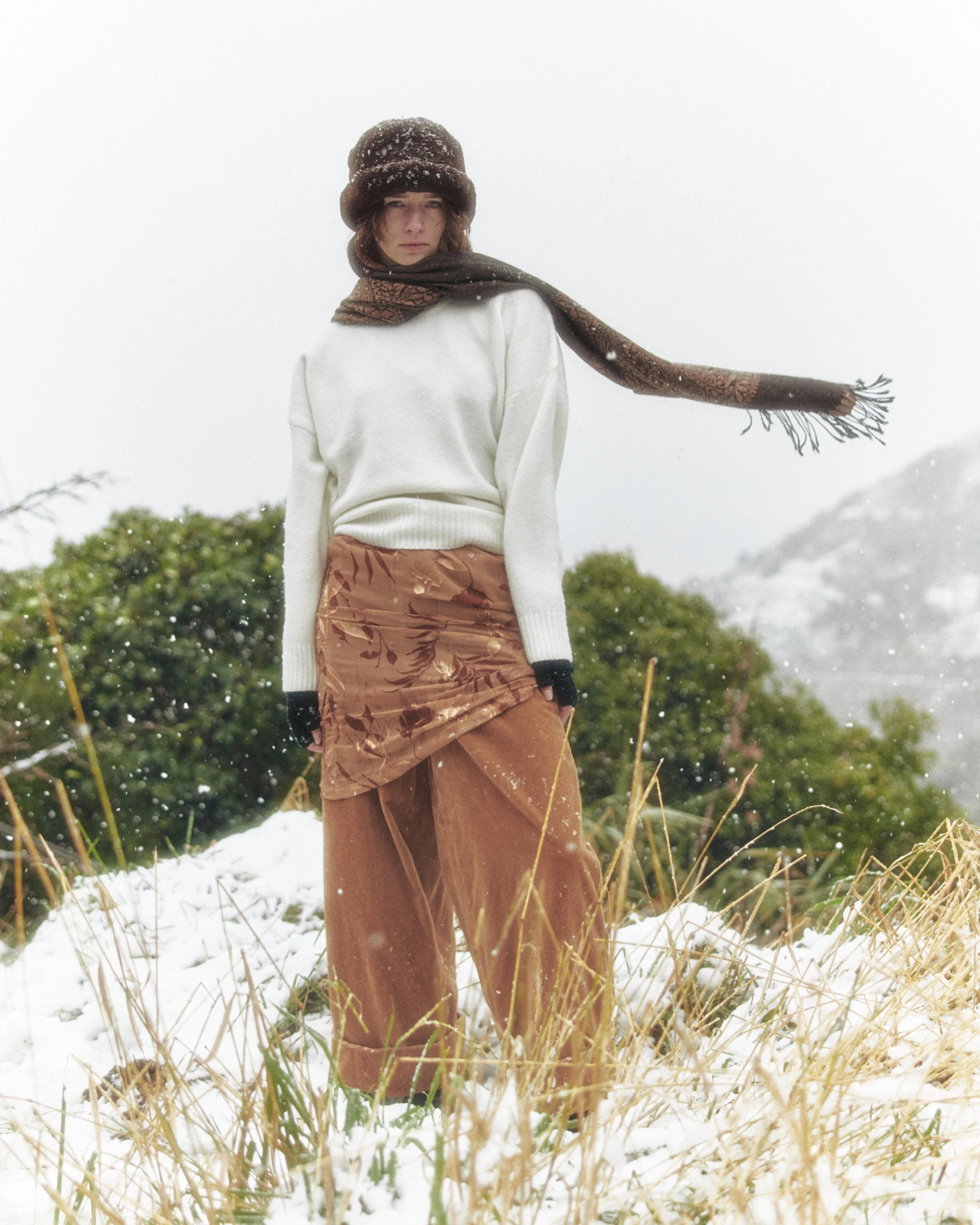 A woman standing outdoors in a snow-covered field with tall grass, wearing a white sweater, brown patterned pants, a dark wool hat with snow, and a brown scarf, with snow falling around her.