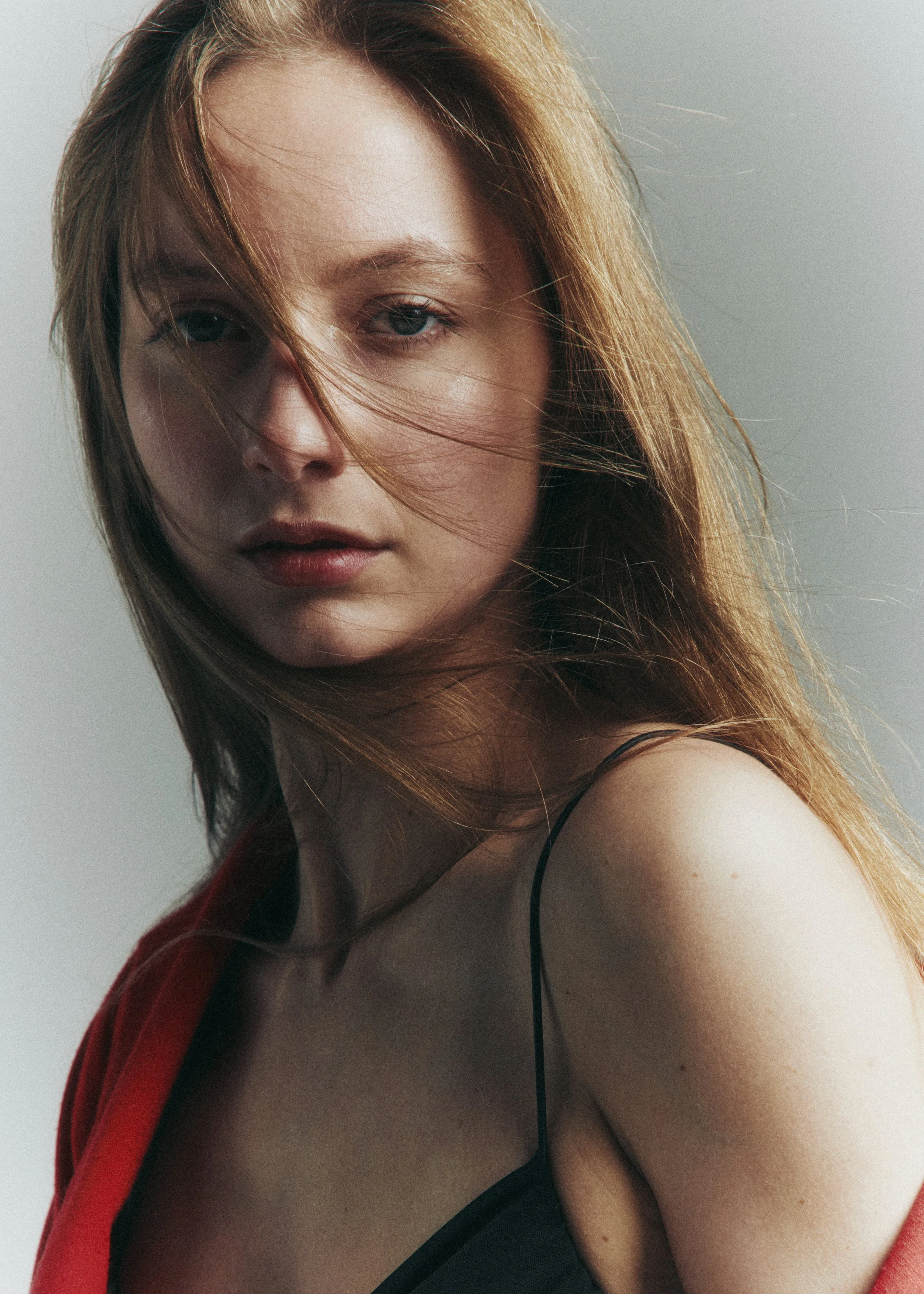 Close-up of a woman with light skin, blue eyes, and long reddish hair, wearing a red and black garment, against a plain gray background.