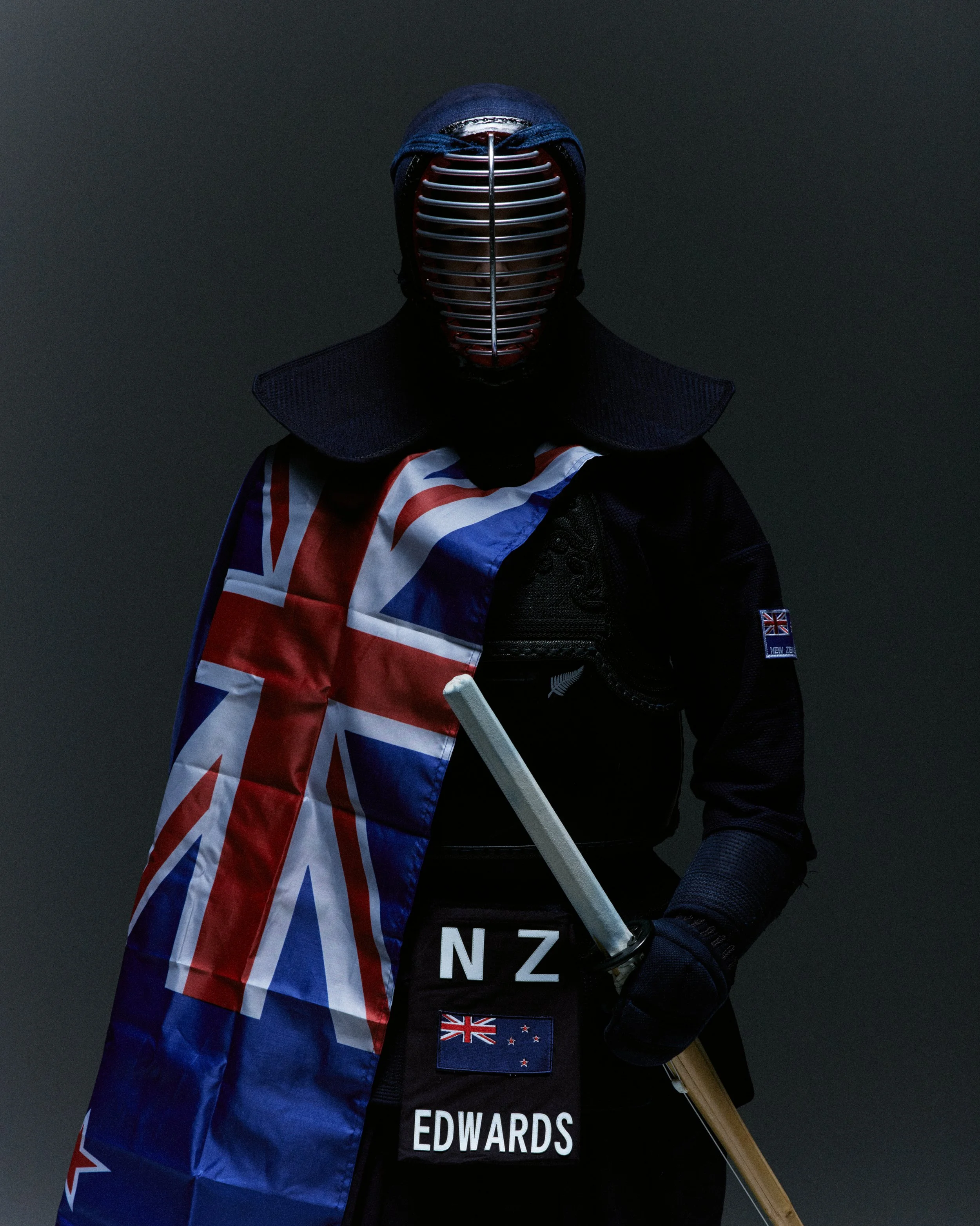 Person wearing a stacked face mask and a kilt, holding a bow, with a New Zealand flag draped over their shoulder, dark background.