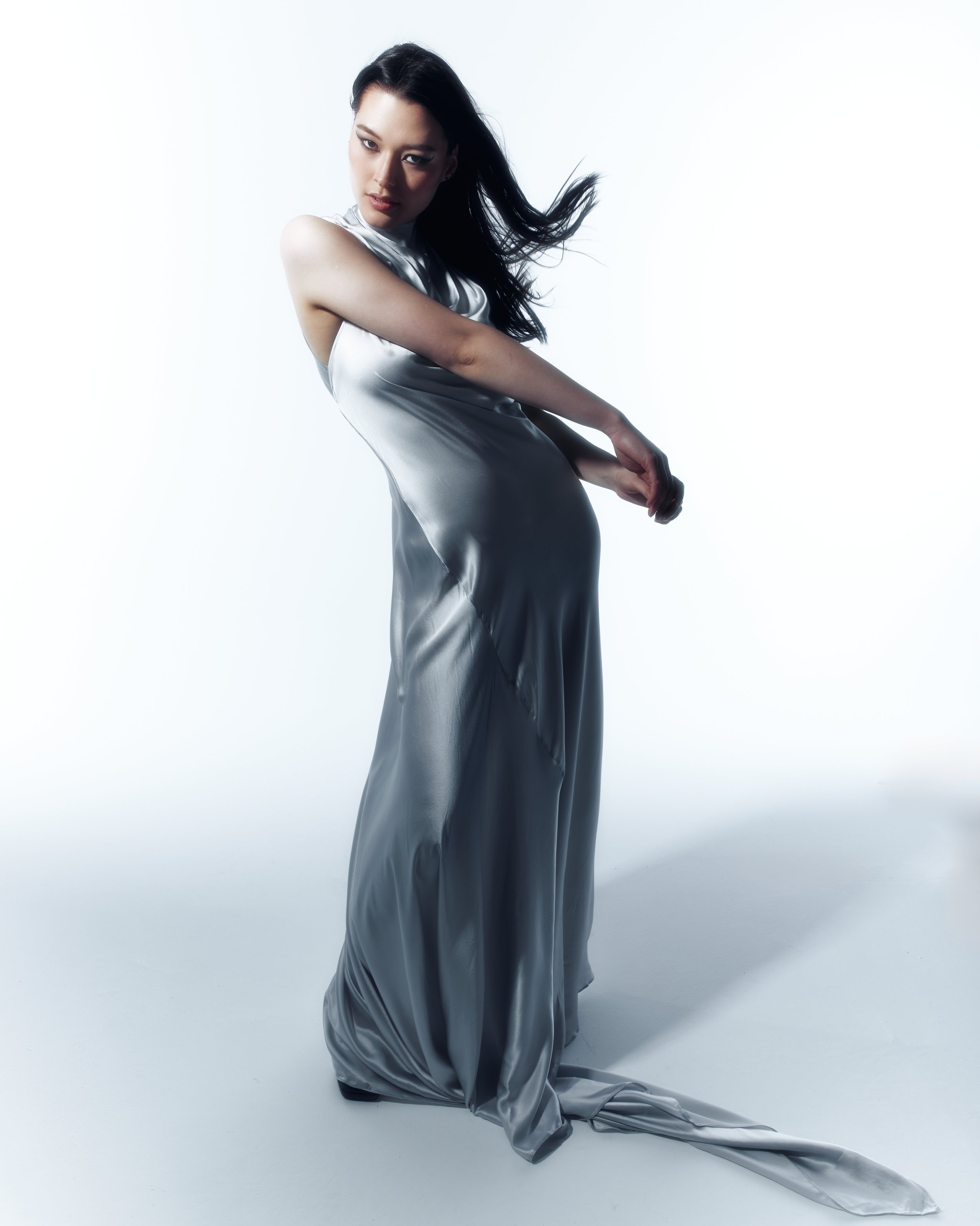 A woman with long, dark hair is wearing a shiny, silver satin dress. She is posing against a white background with a confident expression, one arm bent and the other arm extended downward, with the dress pooling on the floor.
