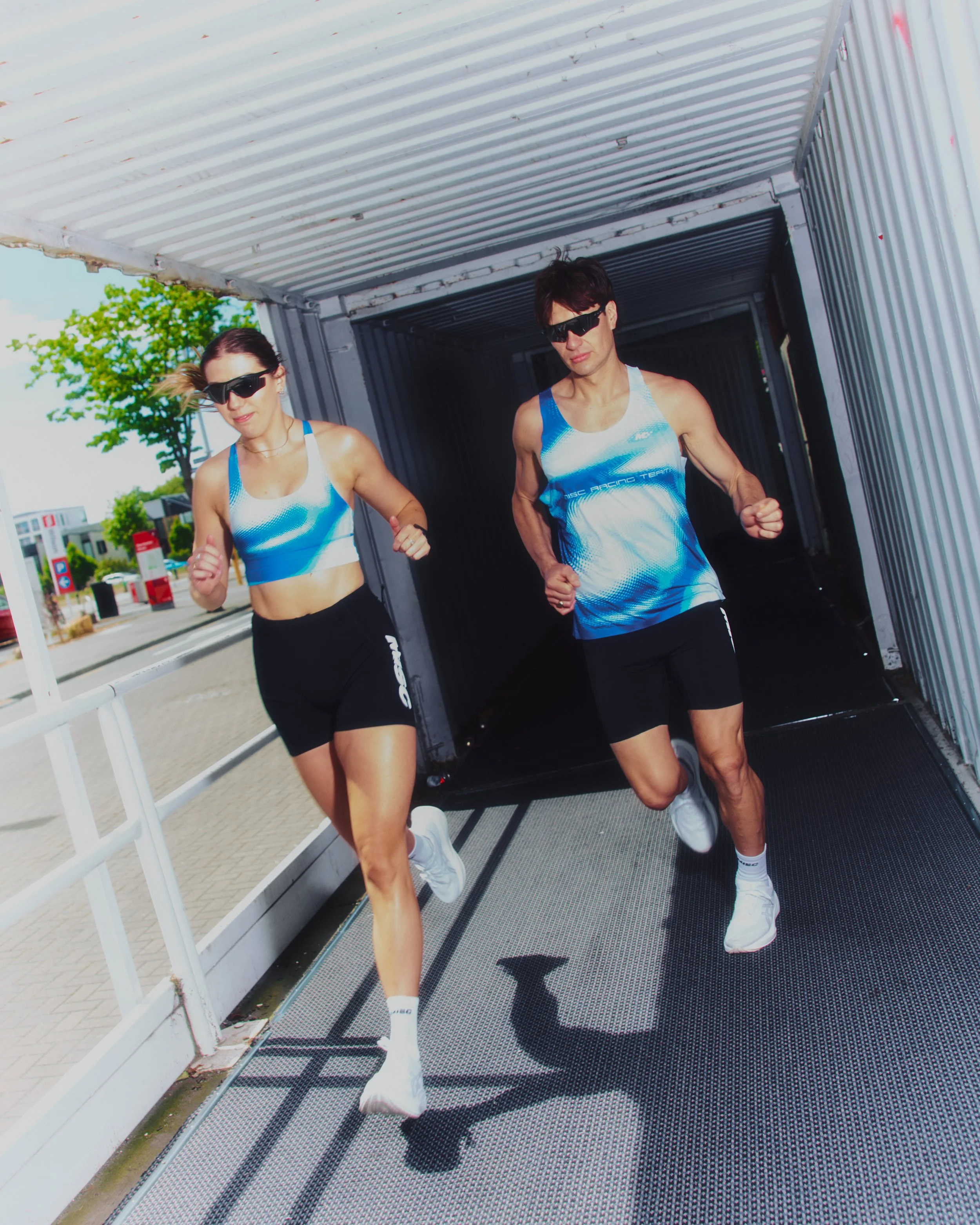 Two runners, a woman and a man, running out of a storage container with a metal floor, during the daytime. Both are wearing blue and white athletic outfits, white shoes, and sunglasses. Trees and a parking lot with cars are visible in the background.