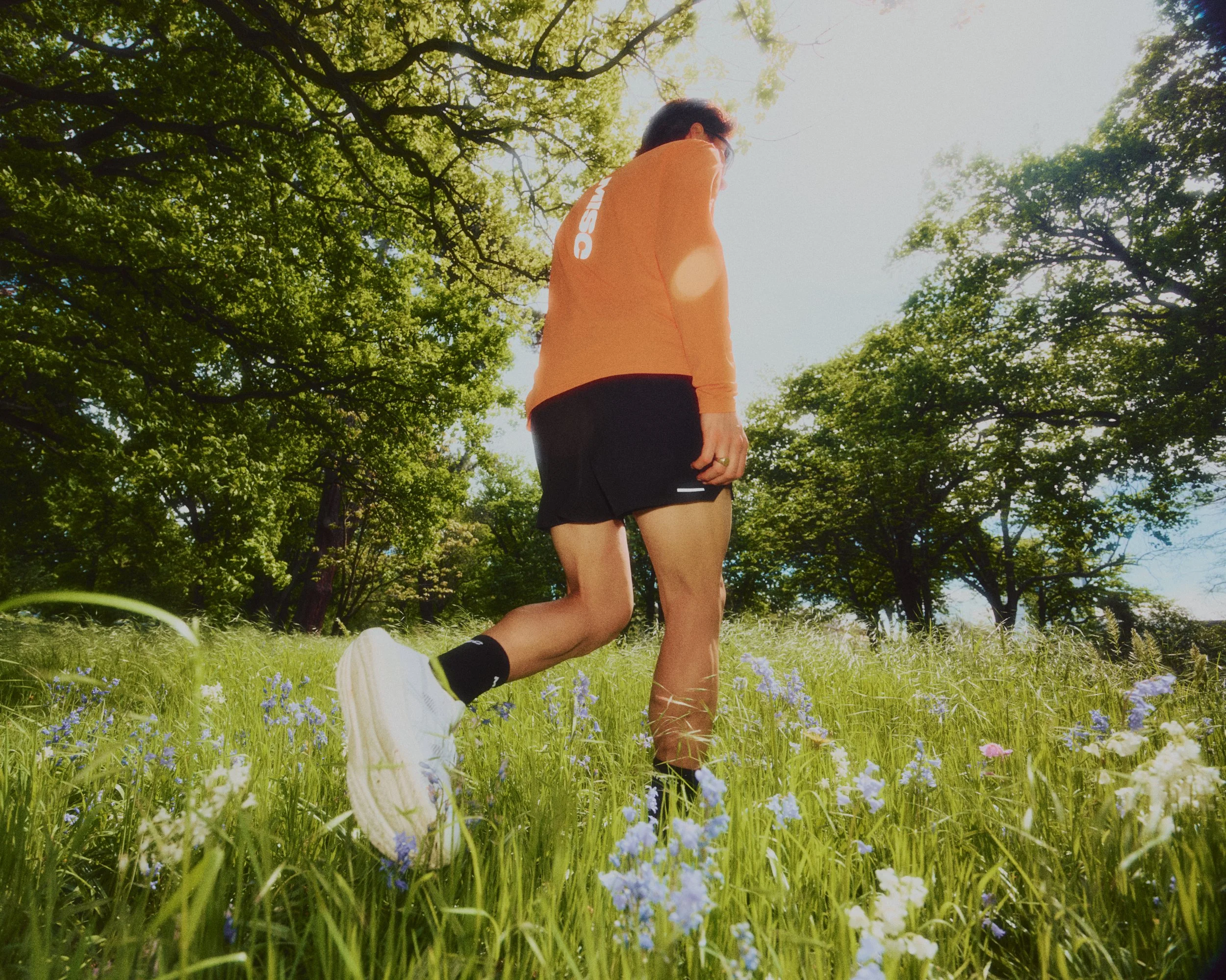 A person jogging outdoors on a grassy field with small purple and white flowers, surrounded by tall trees with green leaves, under bright sunlight.