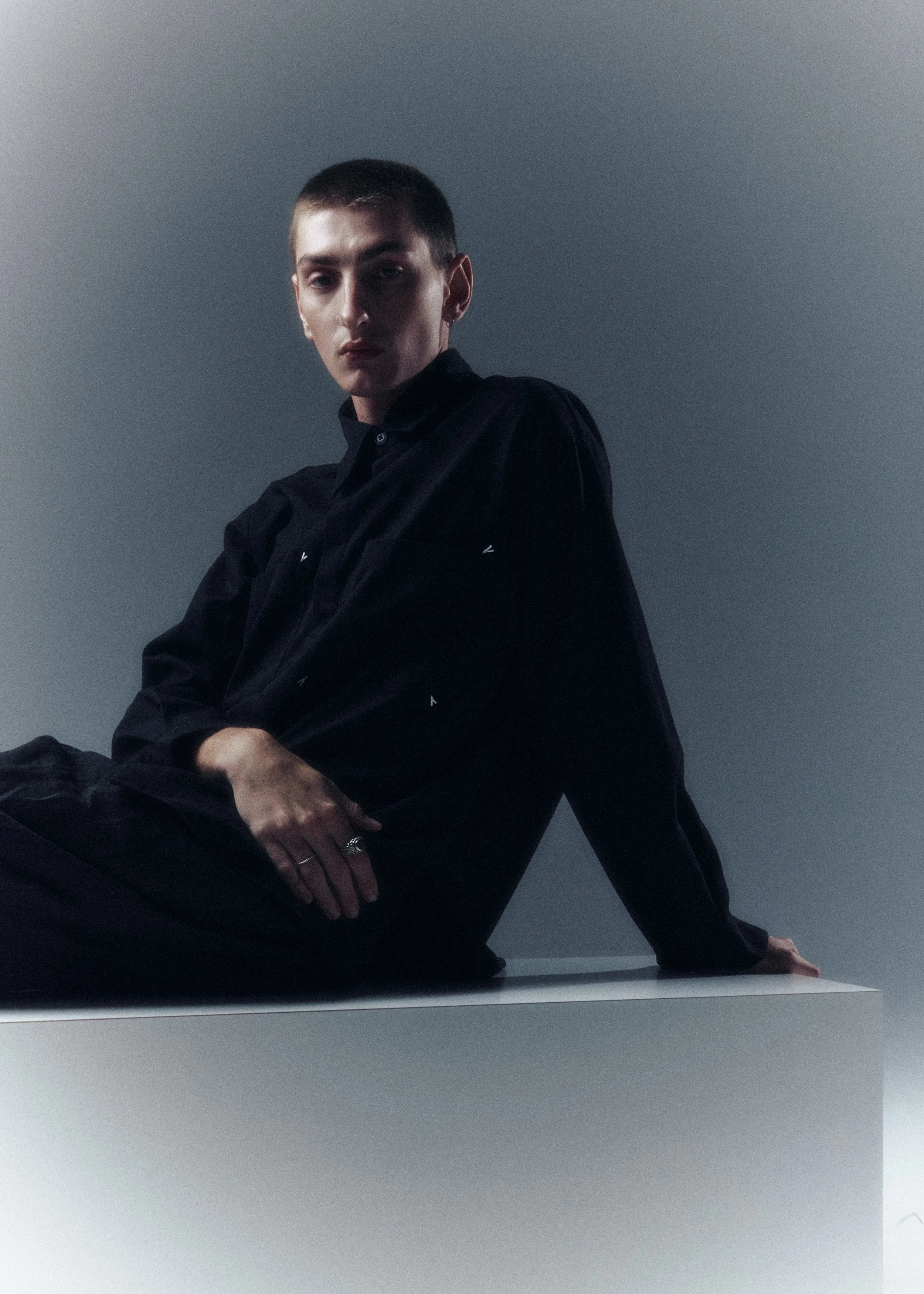A person with short hair and a intense expression, wearing a black shirt, sitting on a table against a plain wall, in a dimly lit environment.