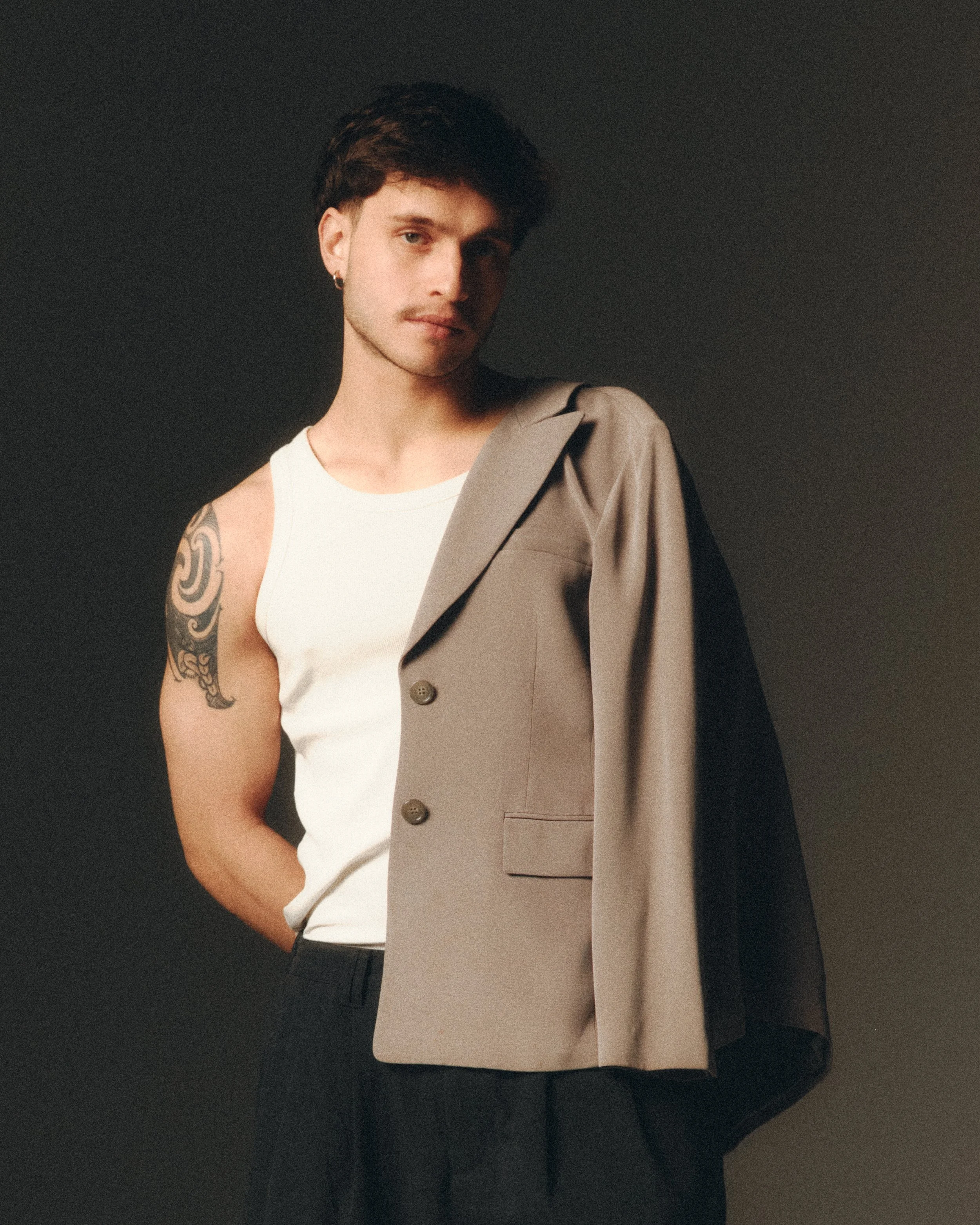 A young man with brown hair, a mustache, and a earring in his left ear is standing against a dark background. He is wearing a white t-shirt and a beige blazer draped over his shoulder, revealing a tattoo on his left upper arm.