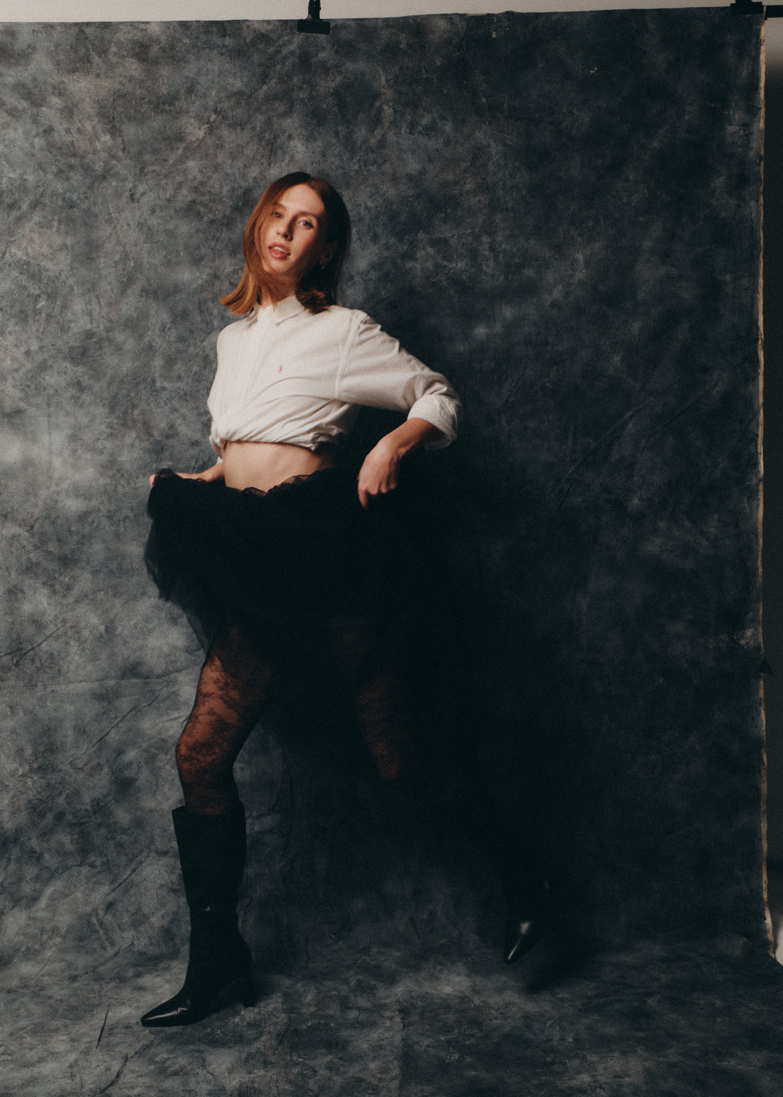 A woman with shoulder-length reddish-brown hair posing against a dark, concrete-textured backdrop, wearing a beige cropped shirt, a black sheer skirt, patterned tights, and black pointed-toe heels.