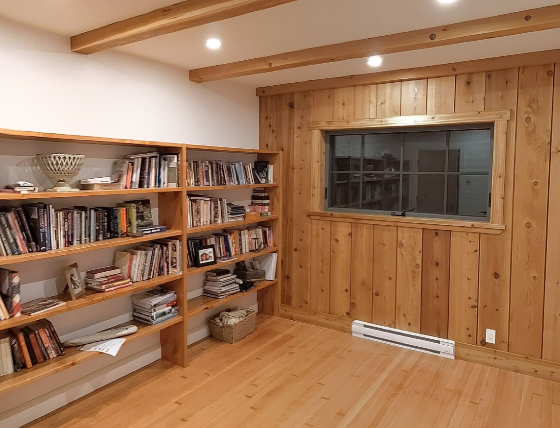 A cozy room with wooden walls, a large window, and a wooden bookshelf filled with books.
