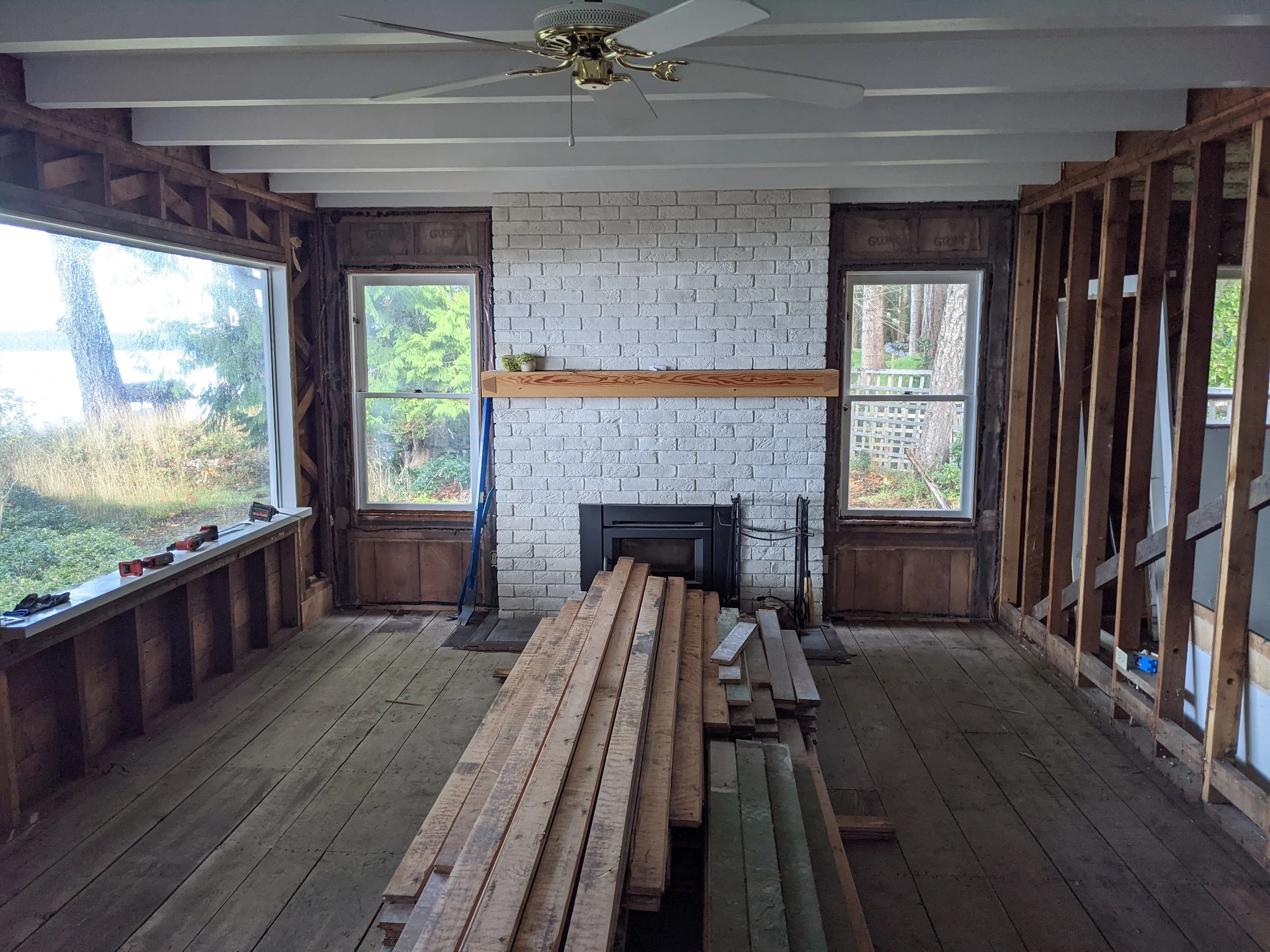 Room under construction with exposed wooden framing, a white brick chimney, large windows showing trees outside, and construction tools and lumber on the floor.