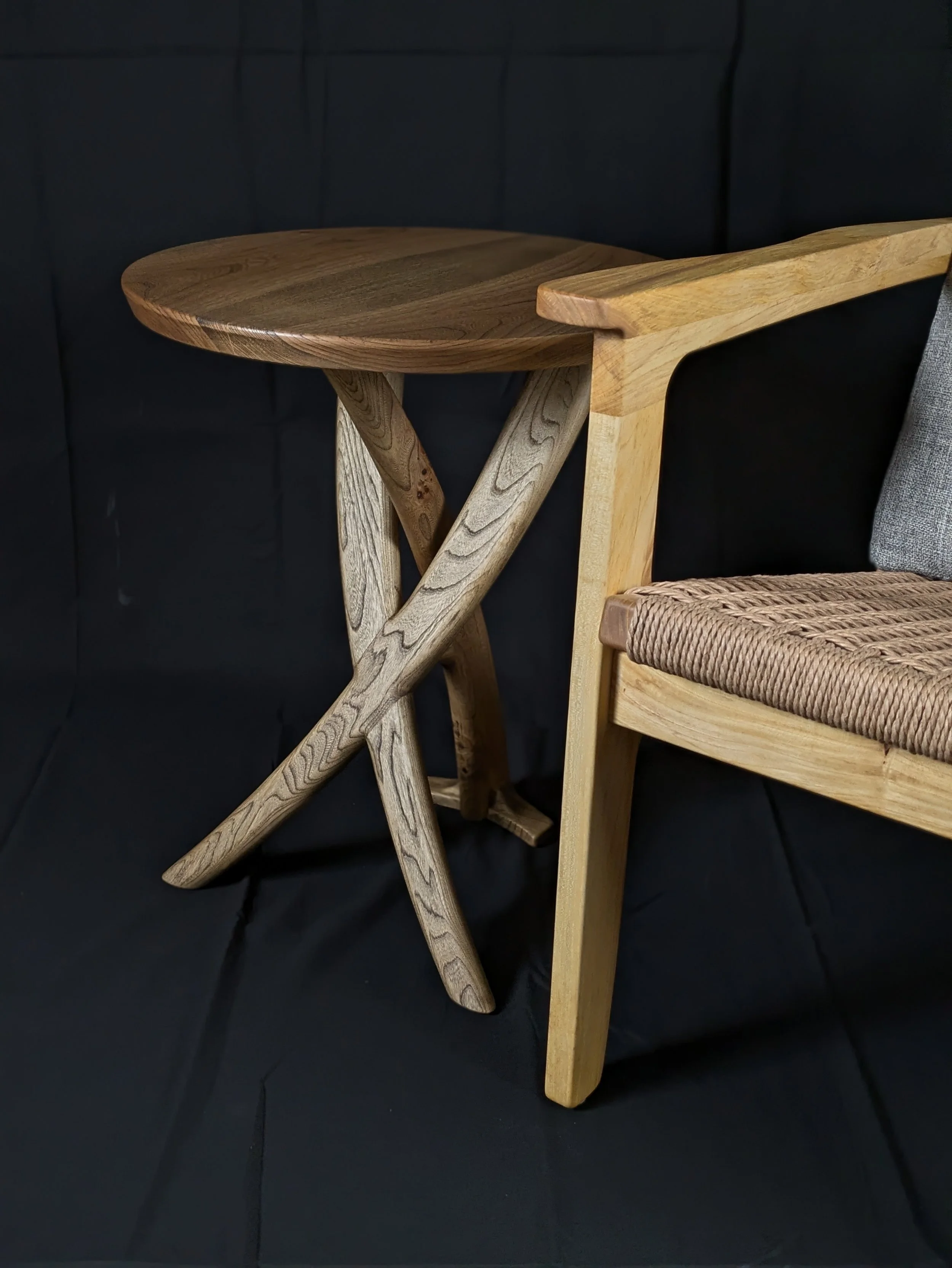 A light-colored wooden side table with a rounded tabletop and an X-shaped support structure, placed against a black background, next to a section of a woven textured chair.