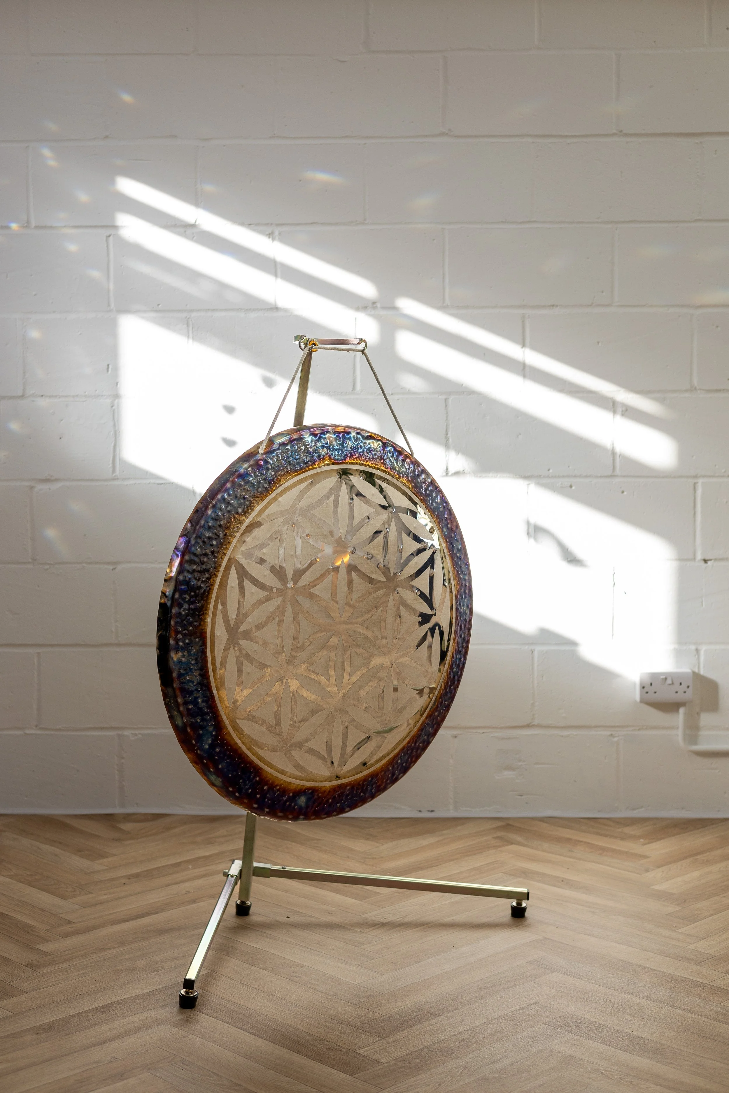HOUSE OF MOON A large gong with geometric flower pattern, supported by a metal stand, set against a white brick wall with sunlight and shadows.