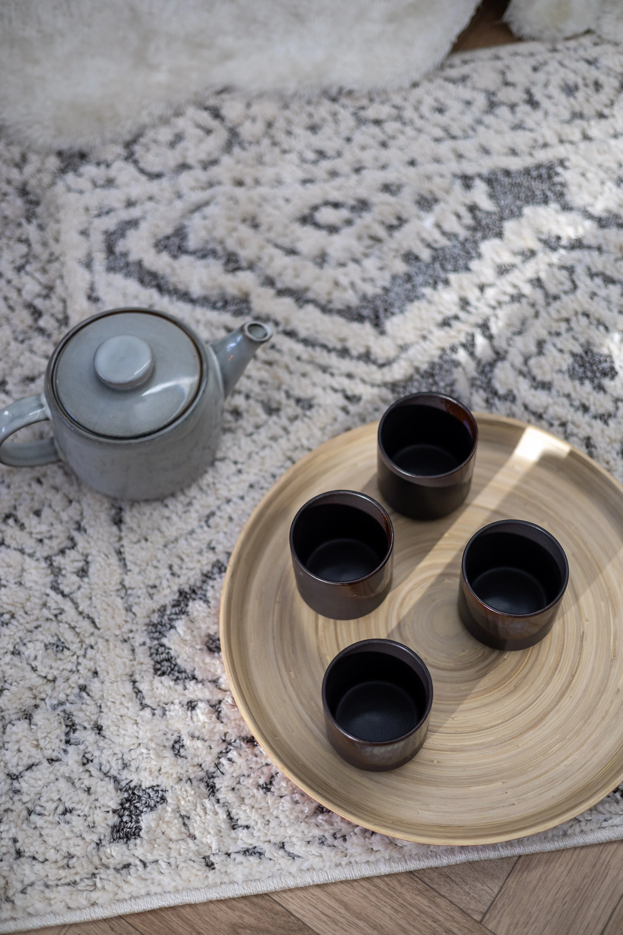 A teapot and four empty cups arranged on a round wooden tray, placed on a fluffy white and gray area rug. HOUSE OF MOON
