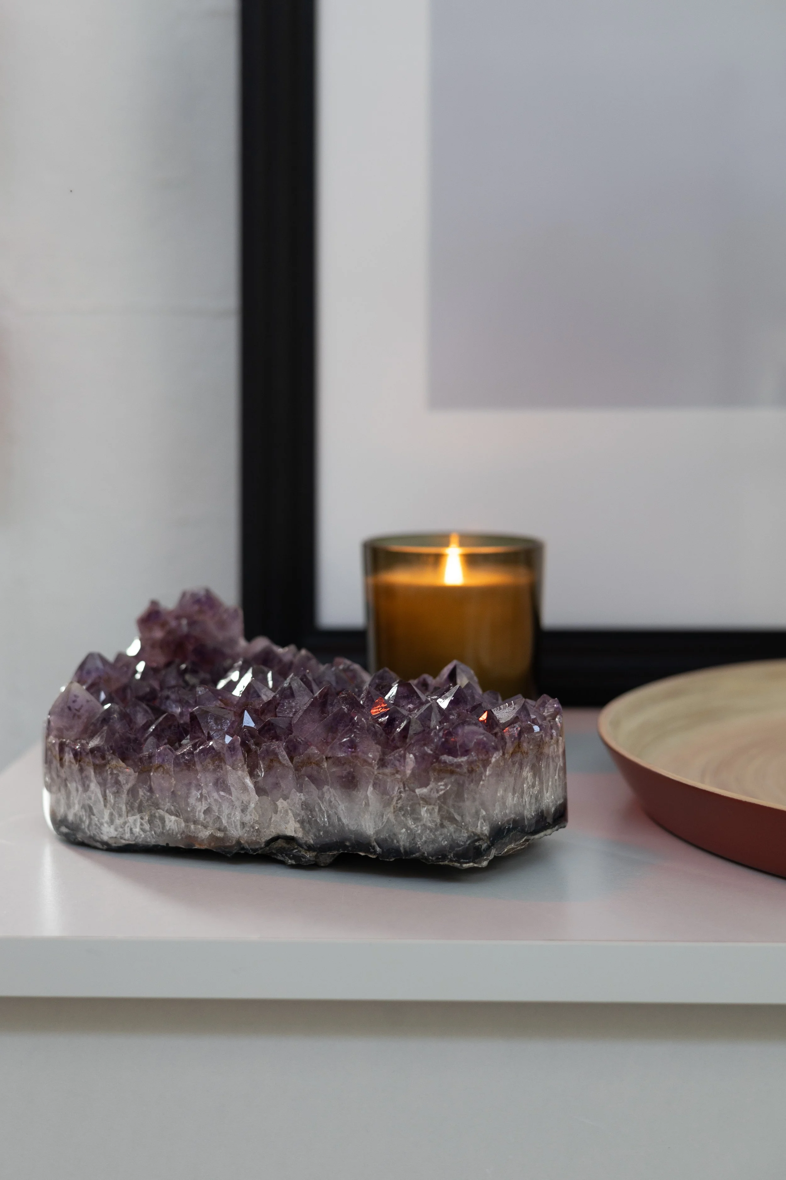 A decorative display on a white surface with a large amethyst geode, a lit candle in a glass holder, a pink ceramic bowl, and a black-framed mirror on the wall. HOUSE OF MOON