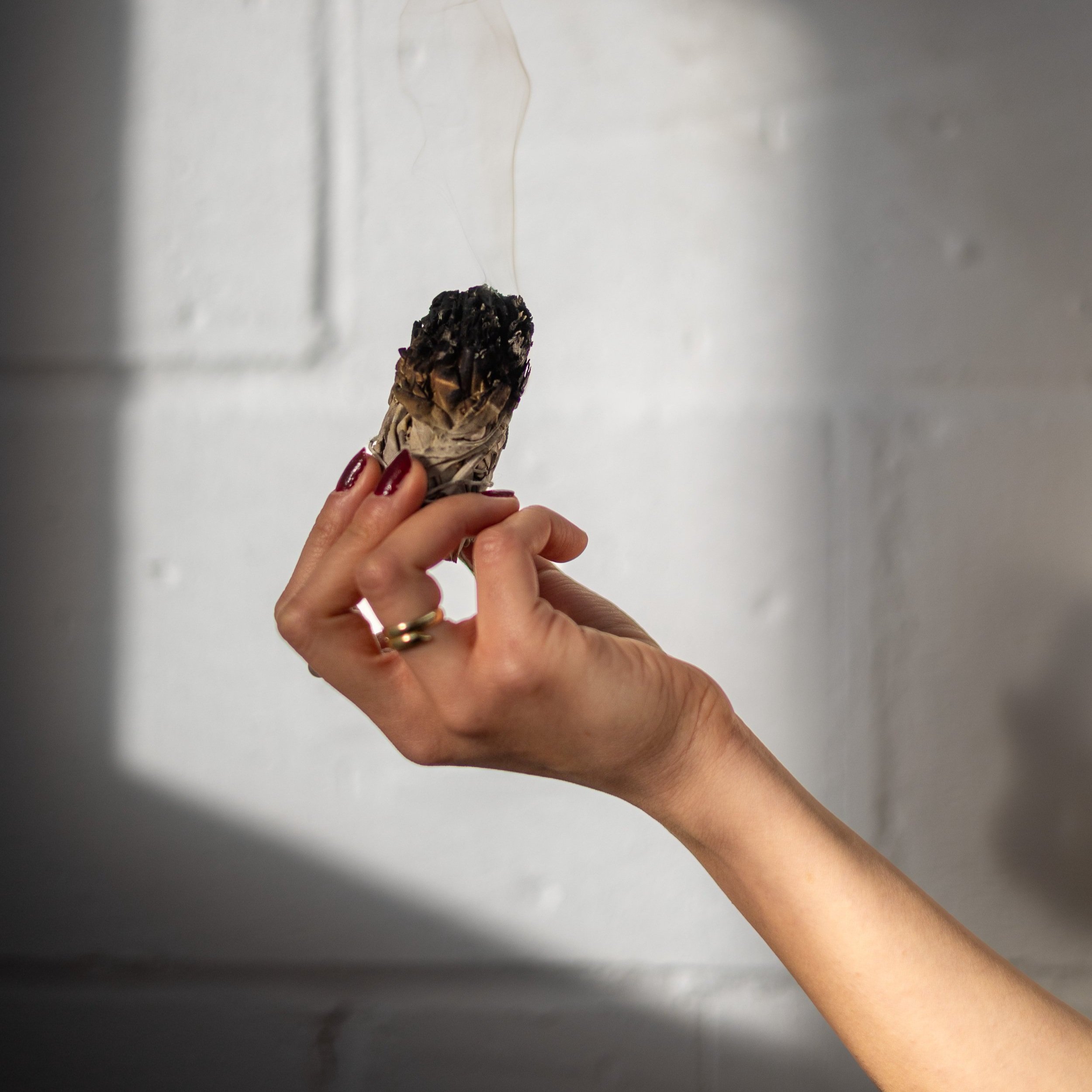 A hand holding sage with smoke rising from the top against a white wall background. HOUSE OF MOON