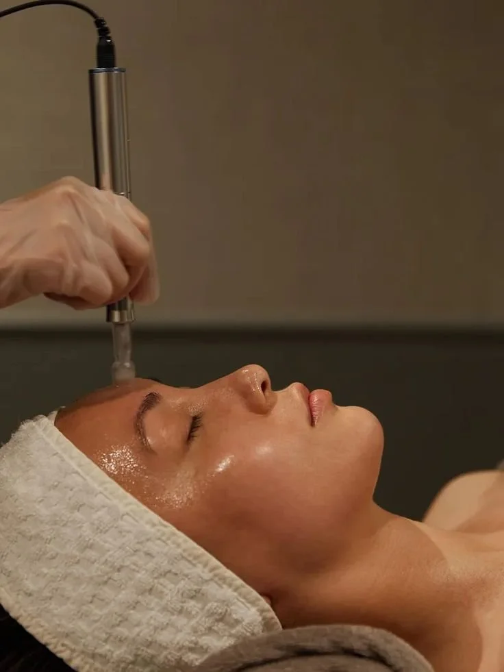 A woman receiving a facial treatment with a handheld device, lying down with a towel wrapped around her head.