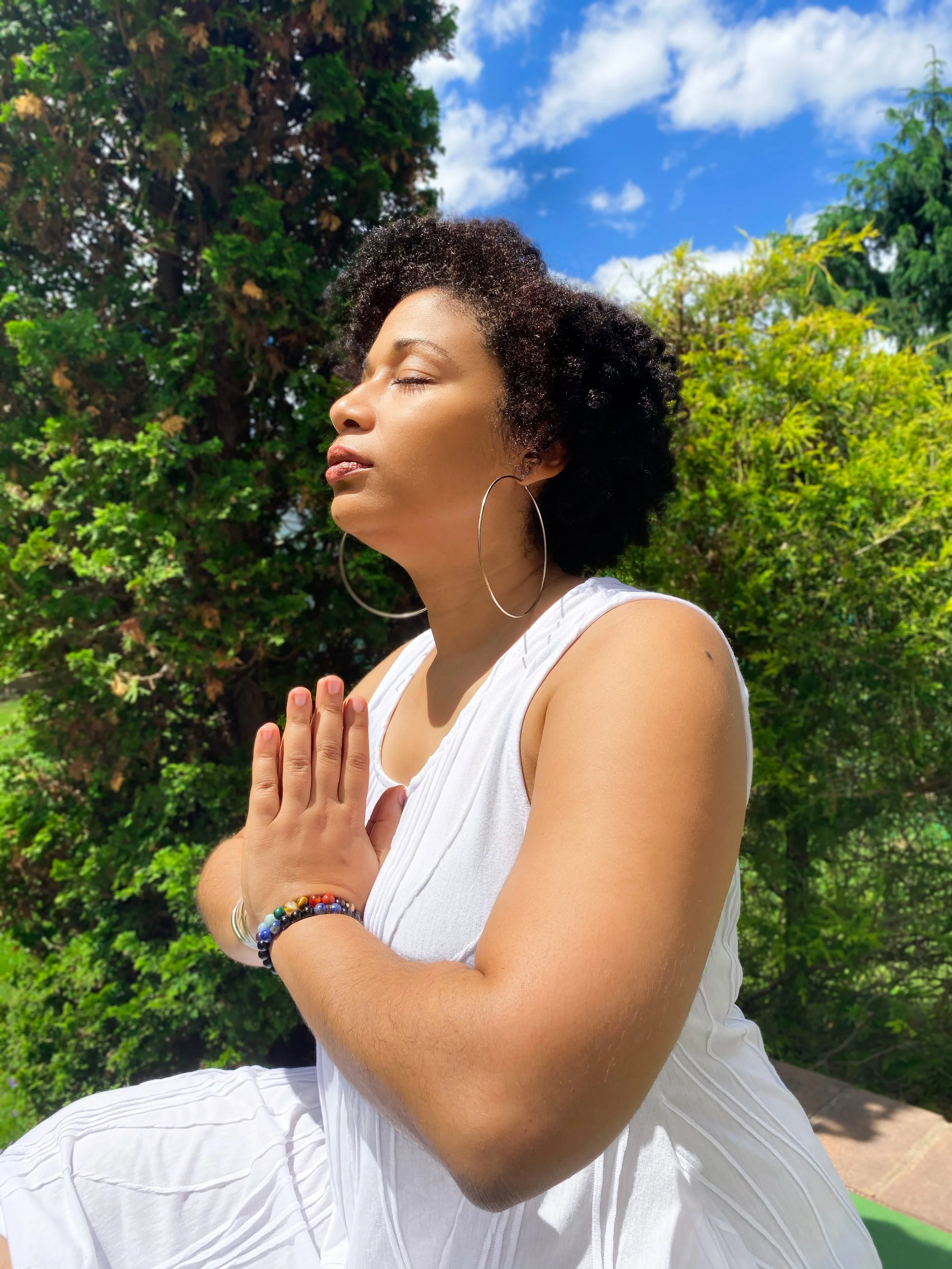 Marjorie is sitting with hands in prayer mode with evergreen trees in background.
