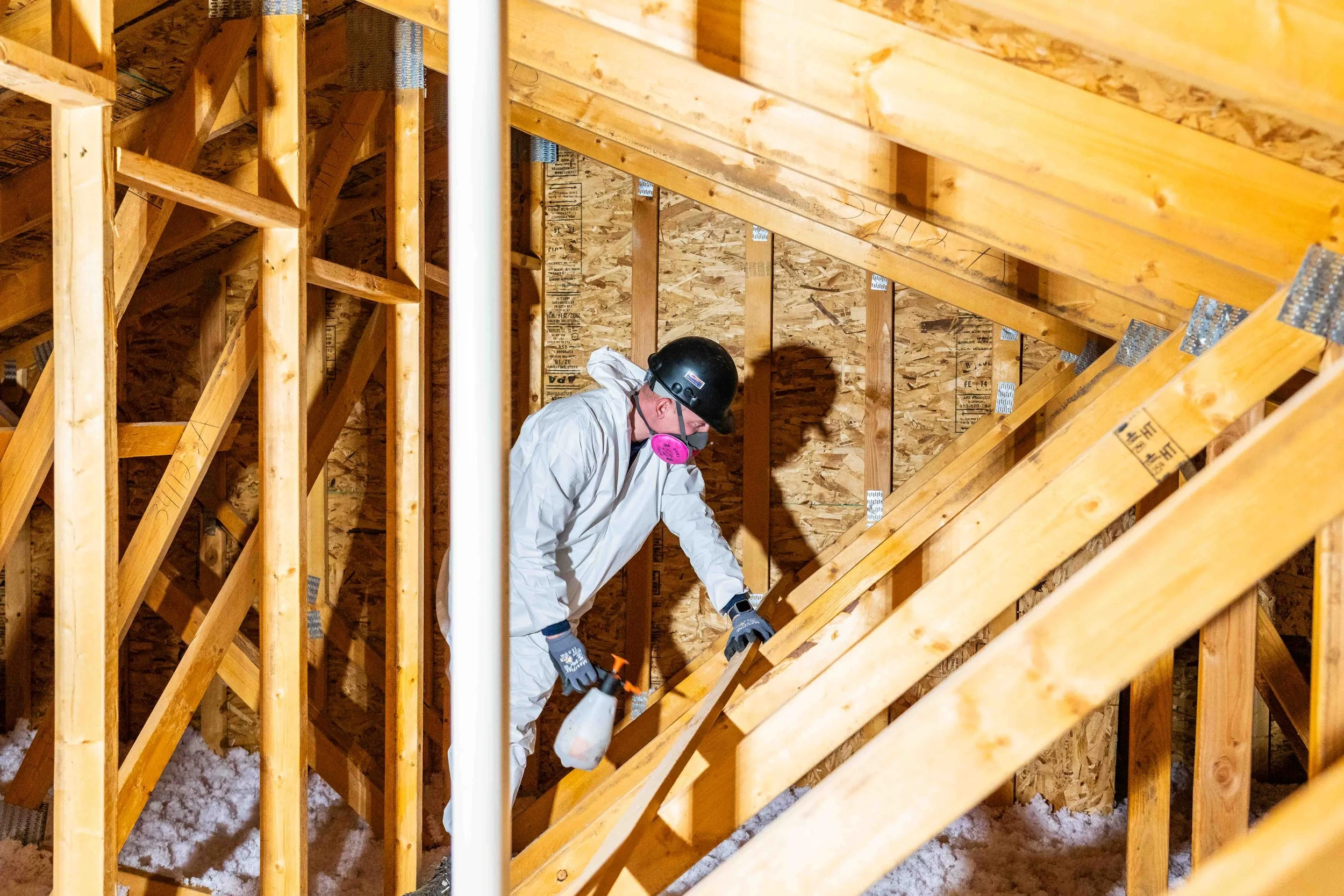 A person in protective gear, including a helmet, gloves, and face mask, is working inside a wooden attic space, using a spray bottle to deodorize the smell left by bats. The unfinished wooden framing and insulation are visible.