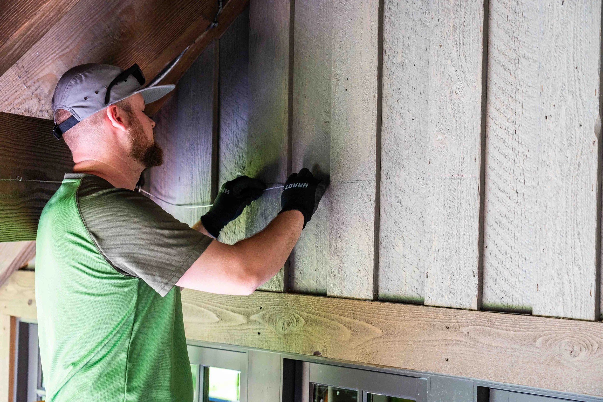 A man wearing a green shirt, black gloves, and a gray cap installing a netting system to prevent birds and bats from entering into an area, using a tool to nail or attach the parts.