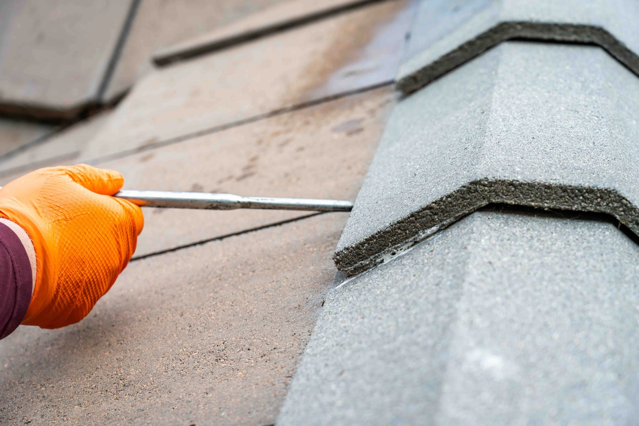 Close-up of a person wearing orange gloves using a metal tool to bat proof a stone tiled roof, with gray tiles visible.