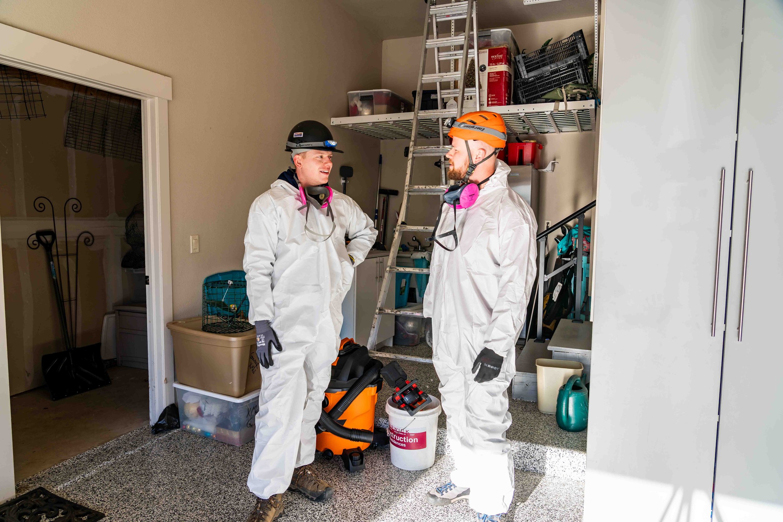Two men in protective gear, including white coveralls, gloves, respirators, and helmets, stand inside a garage, talking and smiling at each other getting ready to go do bat guano remediation and decontamination.
