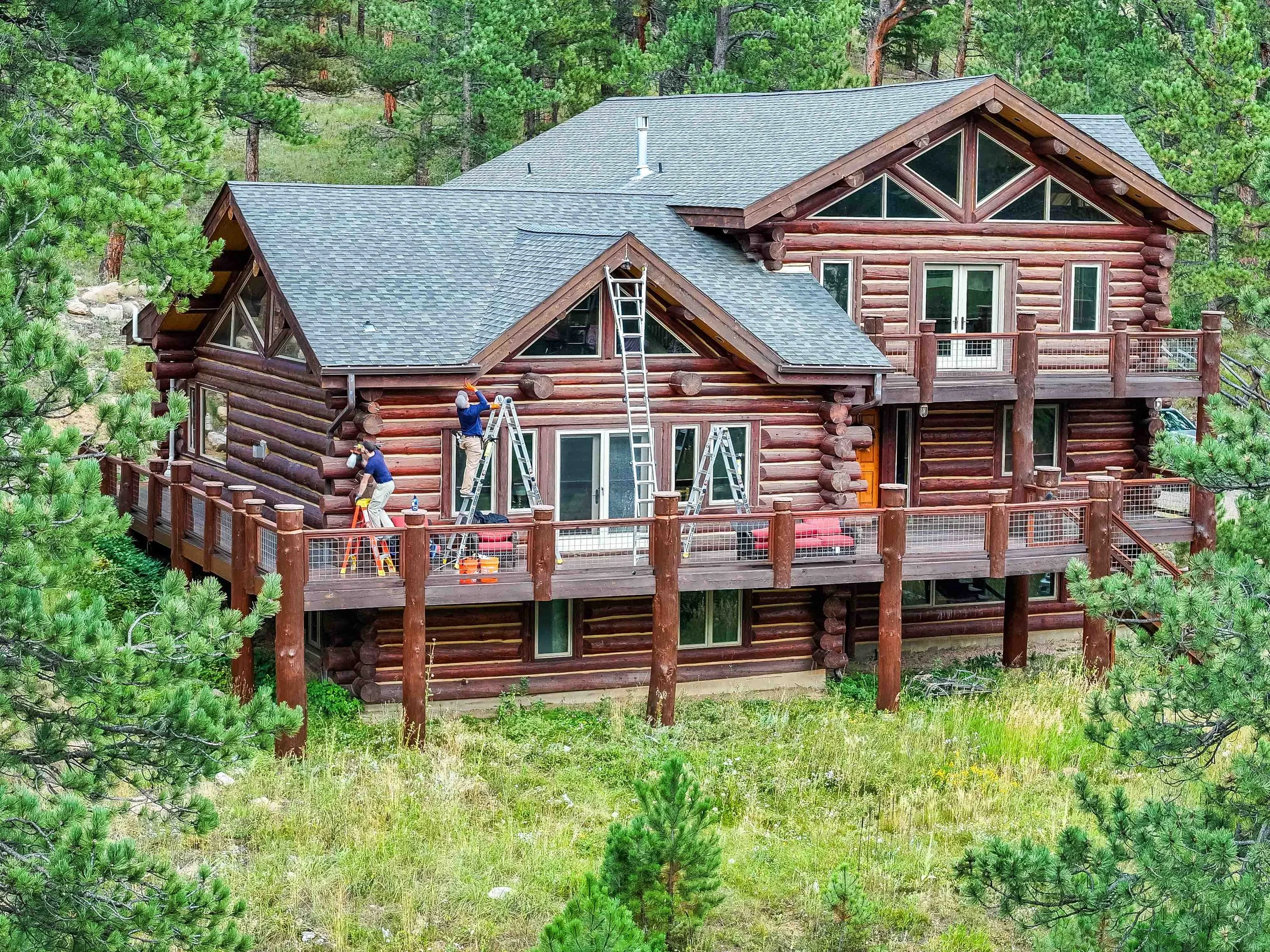Two workers on a large wooden deck attached to a log cabin-style house, performing maintenance and bat proofing, surrounded by pine trees.