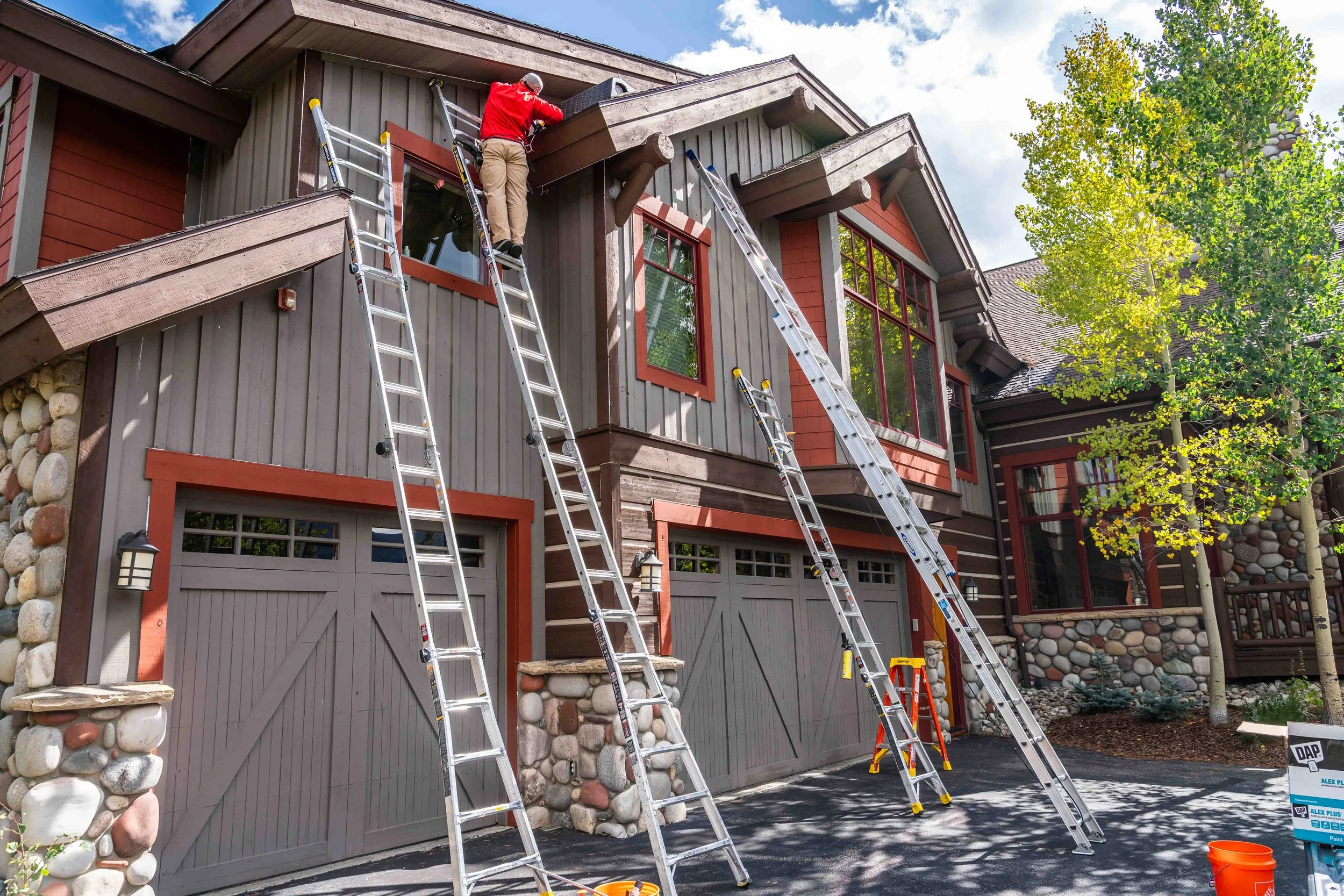 A person in a red shirt and beige pants working on the roof of a multi-story house using ladders, with trees and a clear sky in the background.