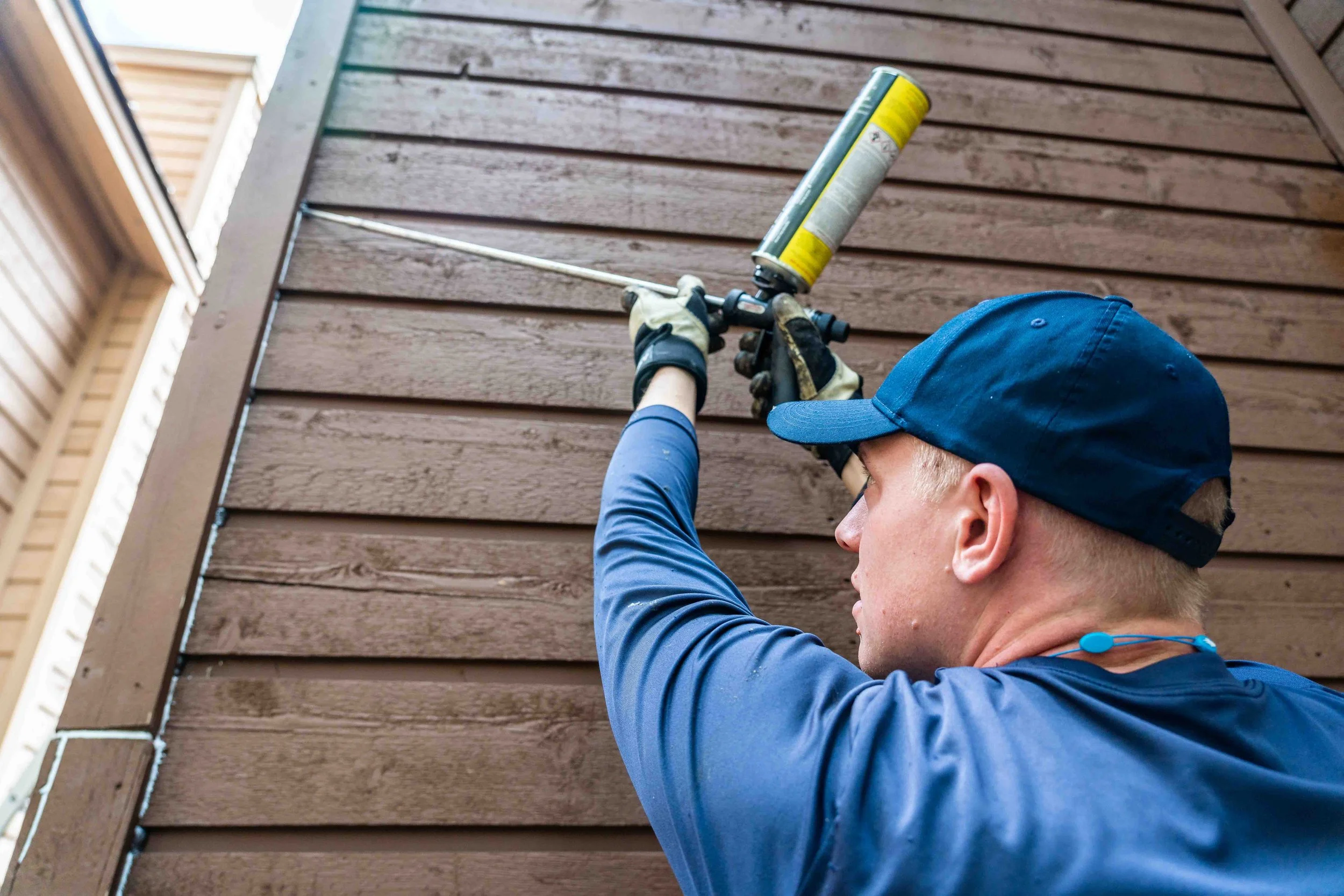 A man wearing a blue cap, blue long-sleeve shirt, and gloves is using a spray foam gun to apply foam insulation to the exterior wall of a house with horizontal wooden siding to prevent bats from entering. 