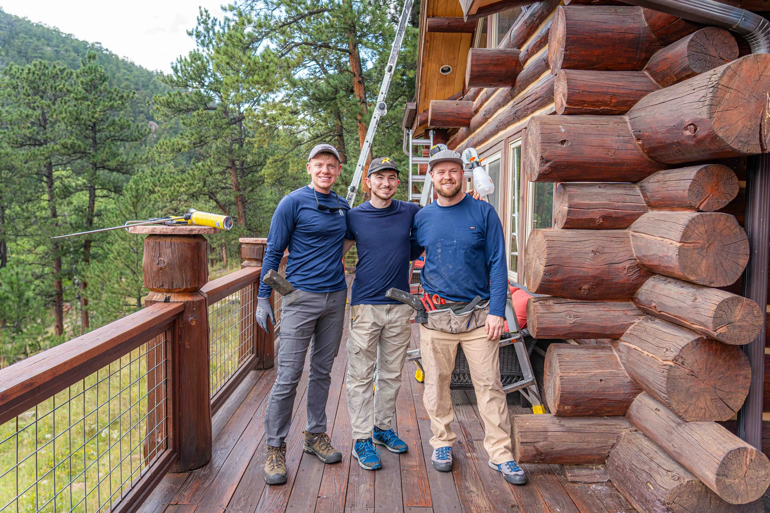 Three men working on a wooden cabin deck, smiling and standing together with tools, surrounded by trees and outdoor scenery.