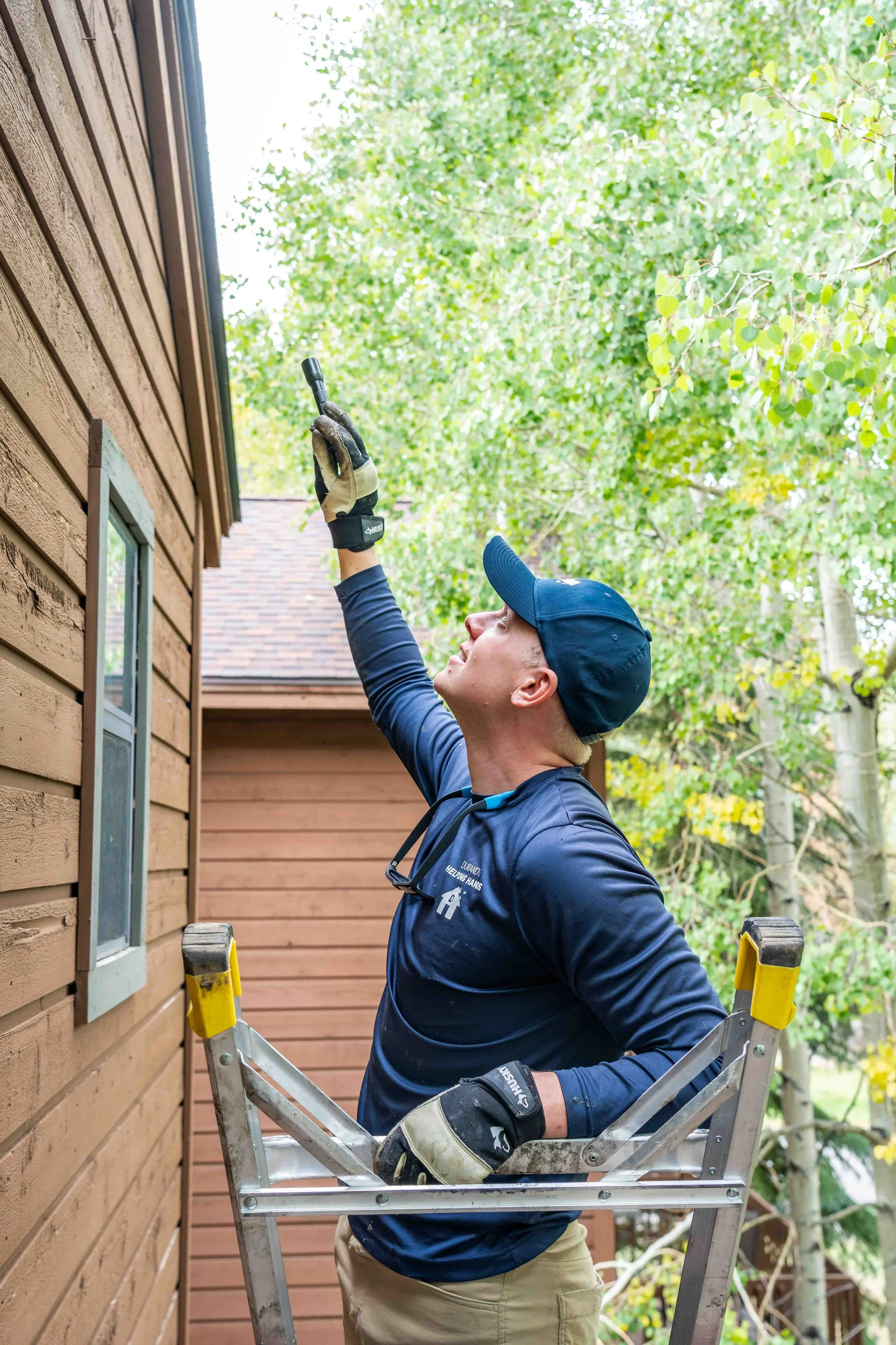 A man standing on a ladder, wearing work gloves and a baseball cap, preforming a Bat Removal Inspection.