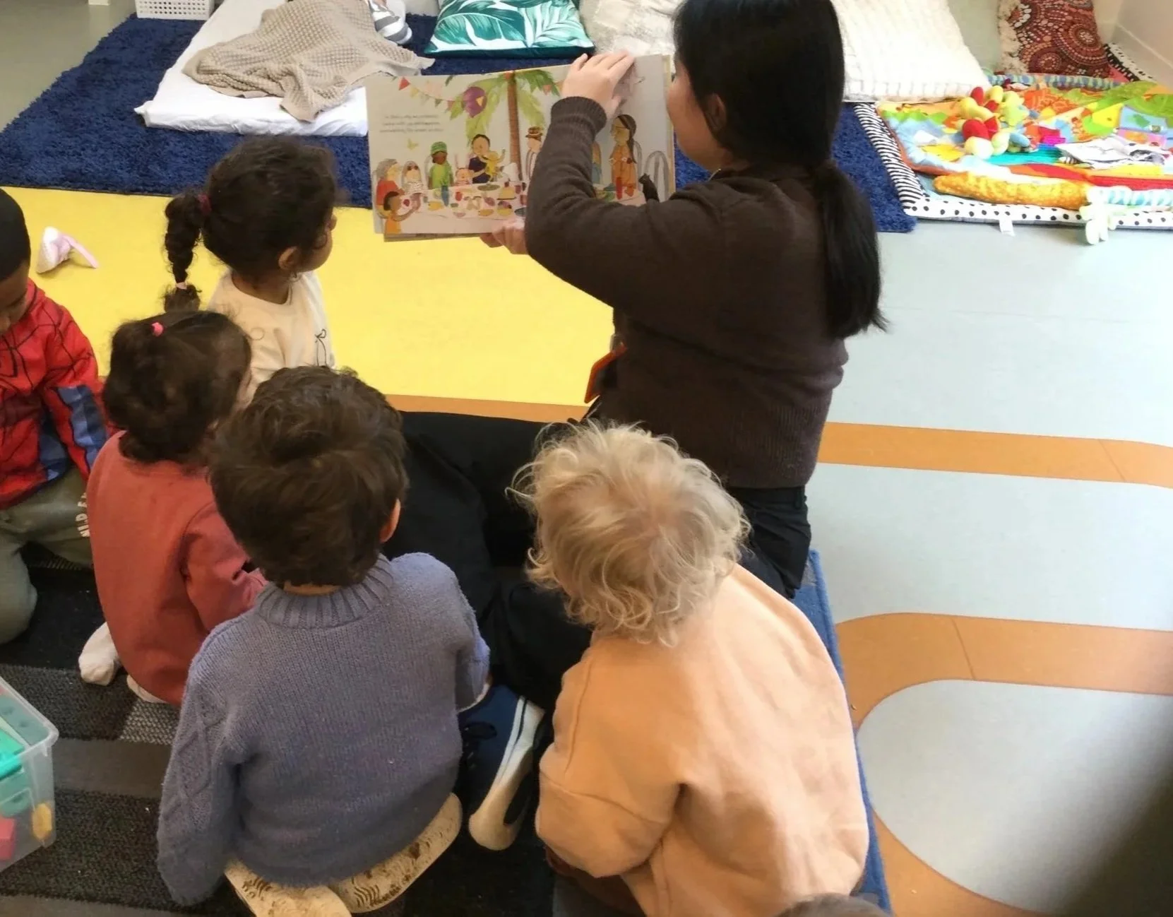 An educator reading a children's picture book to a group of young children sitting on the floor.