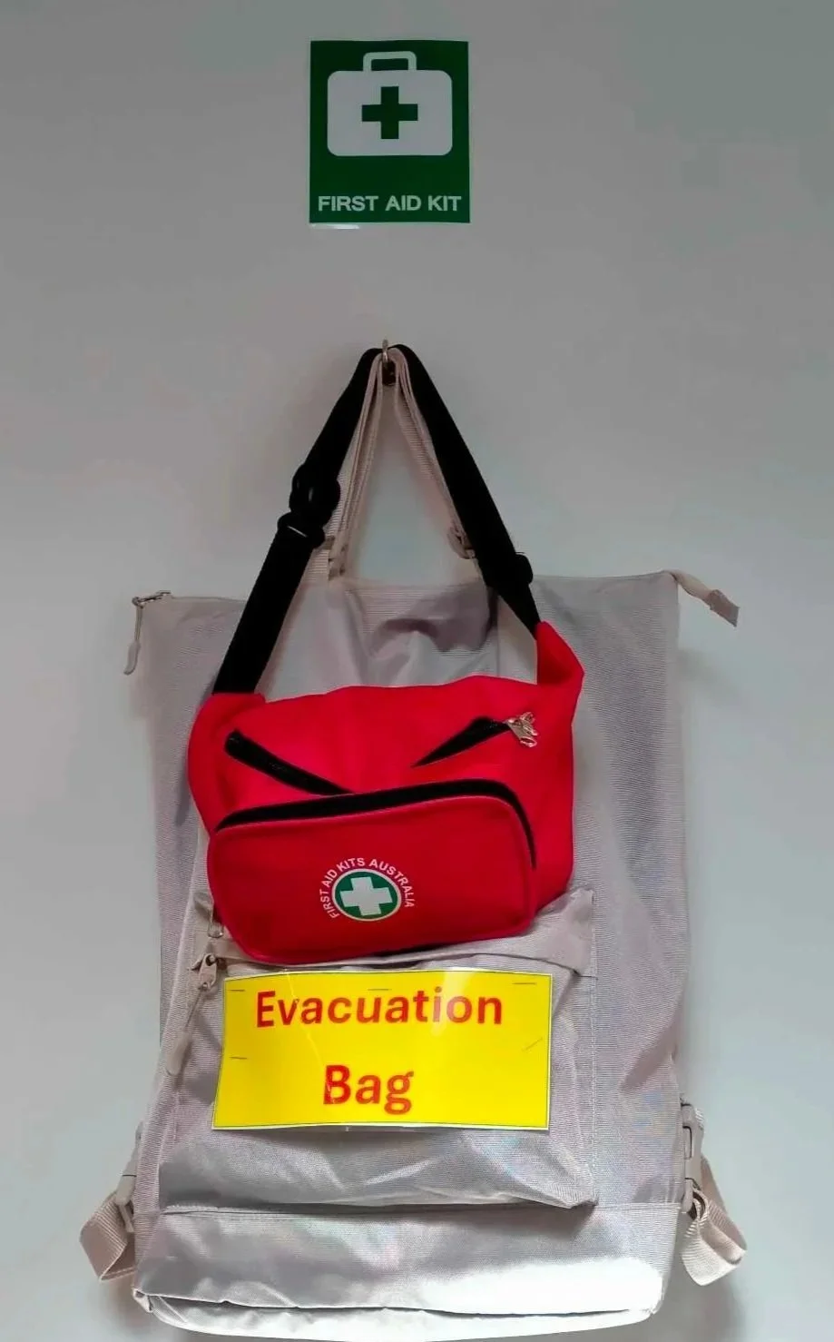 A gray evacuation bag with a red first aid kit bag attached, hang on a wall under an emergency medical green sign, with a yellow sign reading 'Evacuation Bag' in red letters.