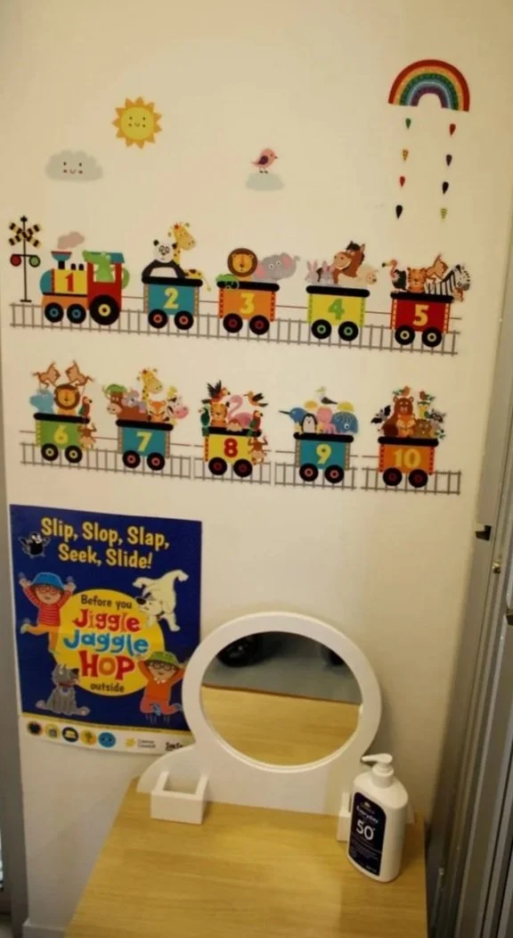 Colorful wall decal of a train with animal characters riding in each car, numbered 1 to 10. A rainbow and weather elements decorate the wall, along with a sign with instructions for a game. A small table with a mirror and hand sanitizer is below the decoration.