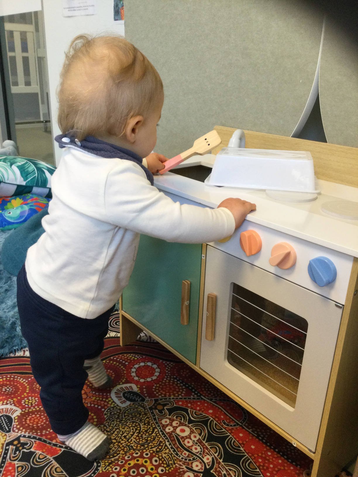 Child playing with a toy kitchen set, holding a wooden spatula, dressed in a white shirt and dark pants, standing on a colorful patterned rug.