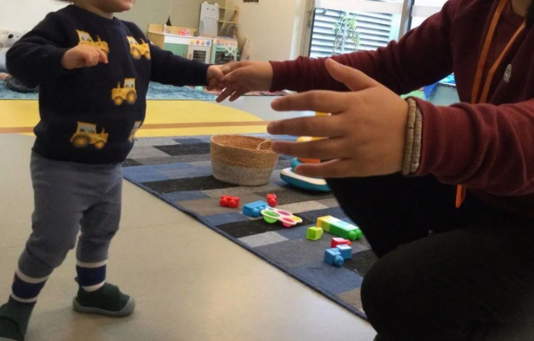 A young child wearing a dark sweater with yellow construction vehicles and gray pants is holding hands with an adult, who is seated on the floor. The adult is wearing a maroon hoodie and has multiple bracelets on their wrist. There are colorful children's toys and building blocks on a rug nearby.