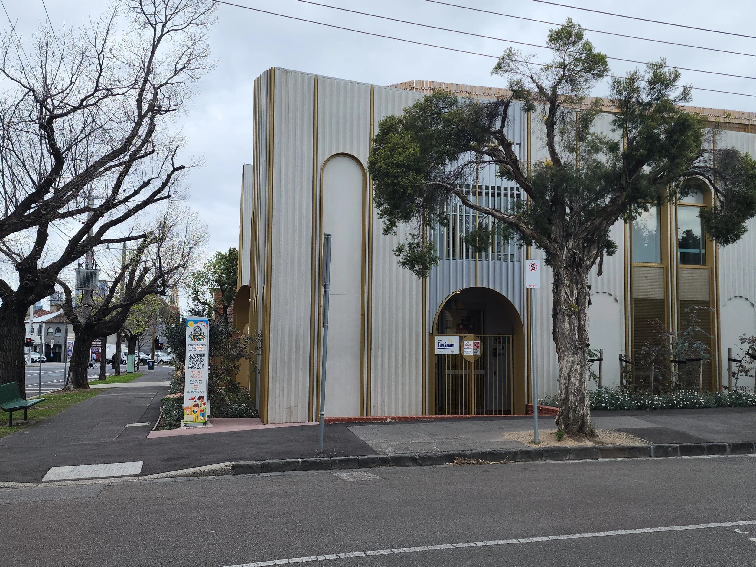 A white building with gold trim and arched windows, partially obscured by trees with bare branches and some green foliage, on a grey overcast day. There is a sidewalk and street in the foreground with a sign and a green bench.