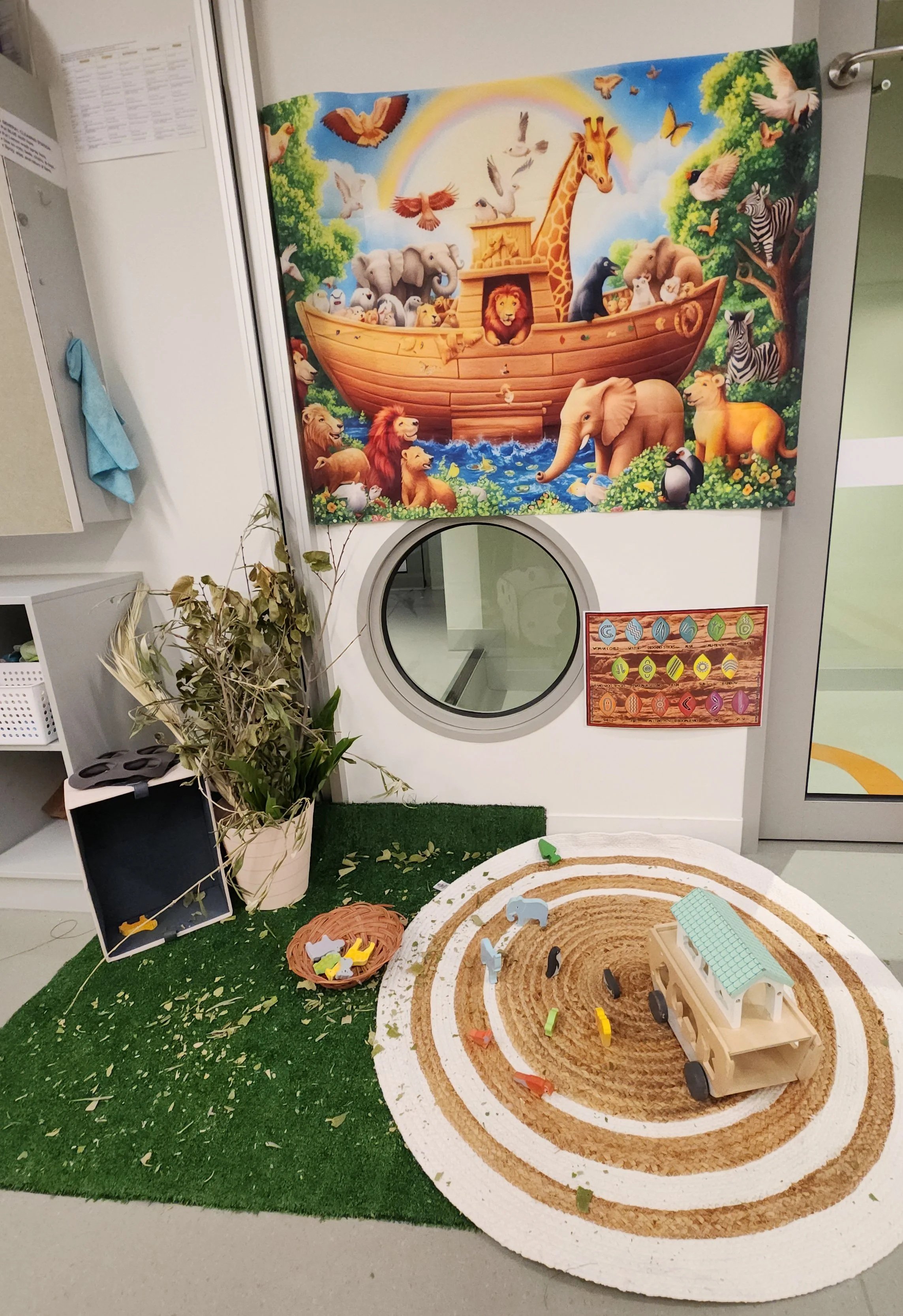 A play area with a forest-themed mural, miniature animal figures, a circular sandpit with toy vehicles, and a small toy house, designed for children's play.