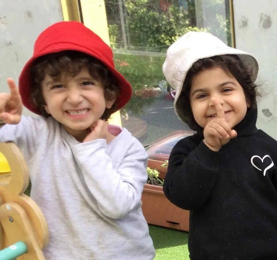At St Mary's Children Centre, Kensington Victoria, two young children with curly hair smiling and wearing hats, one red and one white, making the gesture of "number 1", with a glass window showing outdoor greenery.