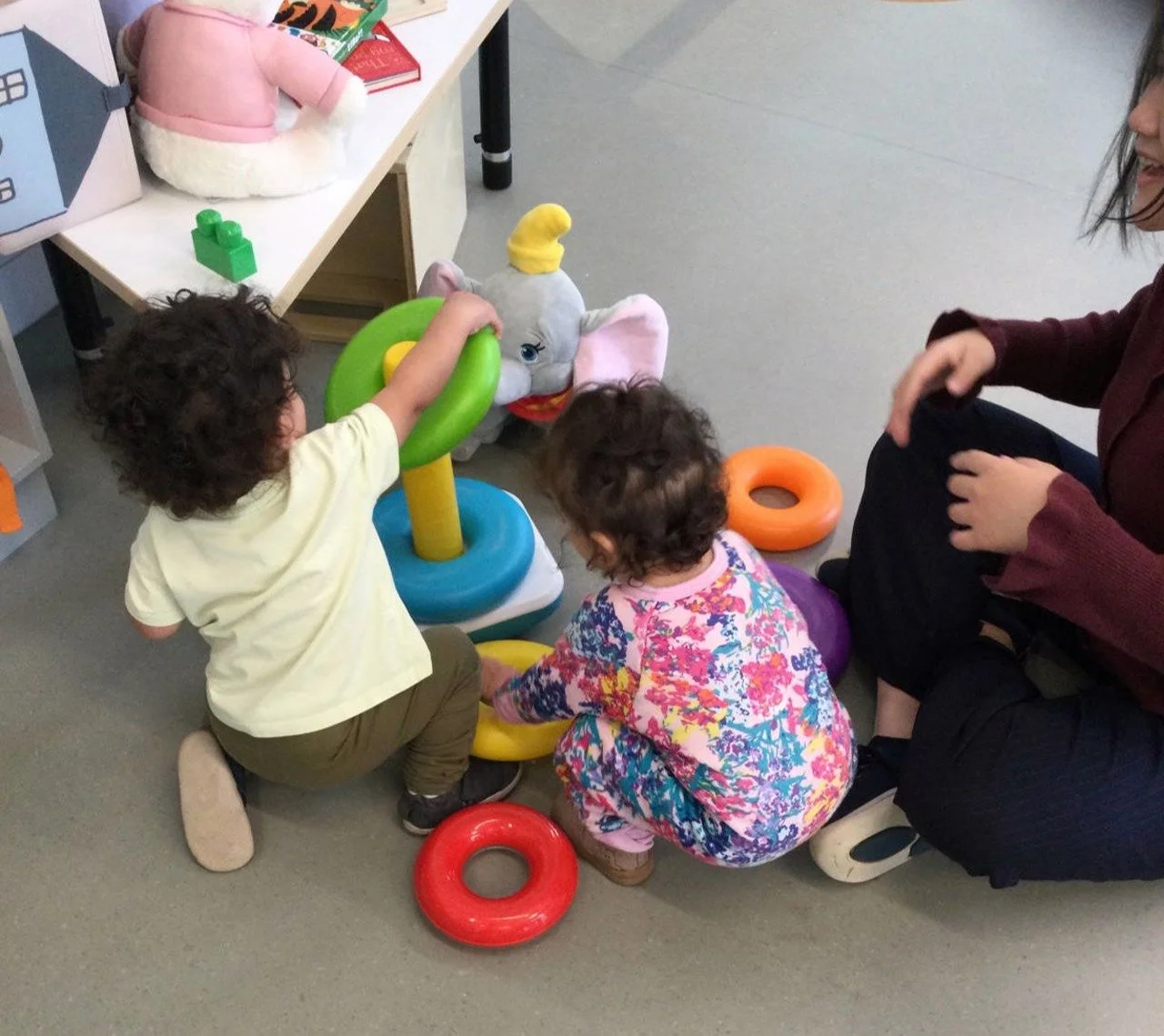 Two children and an adult playing with a plush elephant and colorful rings on the floor. The children are kneeling, and the adult is sitting nearby, engaging with them in a play area.