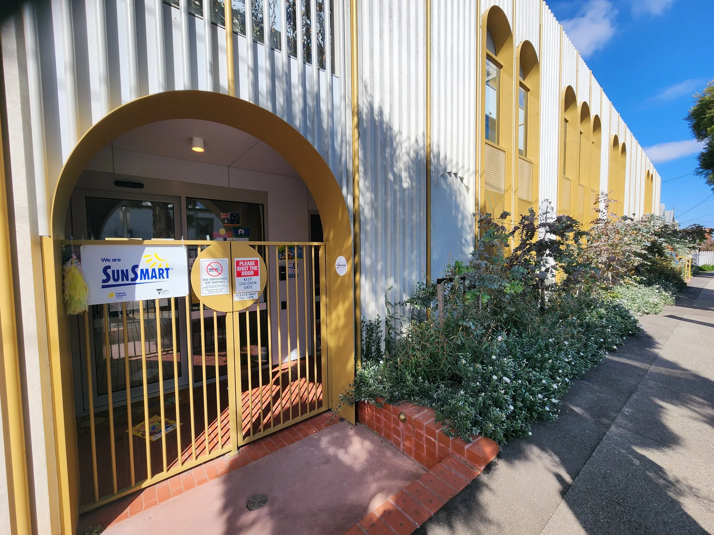 Yellow or golden gate and doorway at the entrance to a building that houses child care centre, with signs indicating no smoking and shutting the door, surrounded by greenery and a sidewalk. Building located at the corner of Epsom Rd and The Ridgeway.