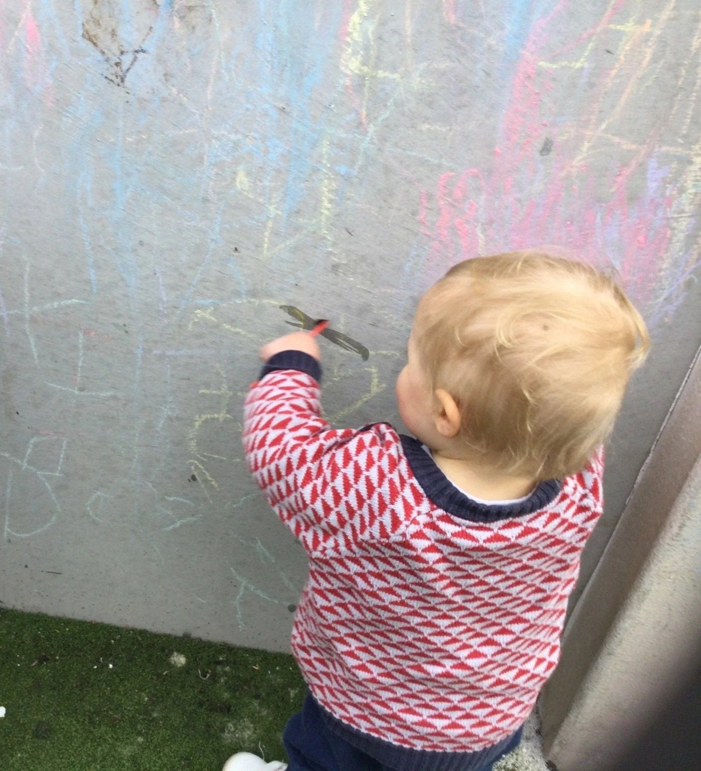 A young child with blonde hair playing with chalk on a gray concrete wall. The child is wearing a red and white patterned sweater and dark pants, facing away from the camera. There are colorful chalk drawings and scribbles on the wall.