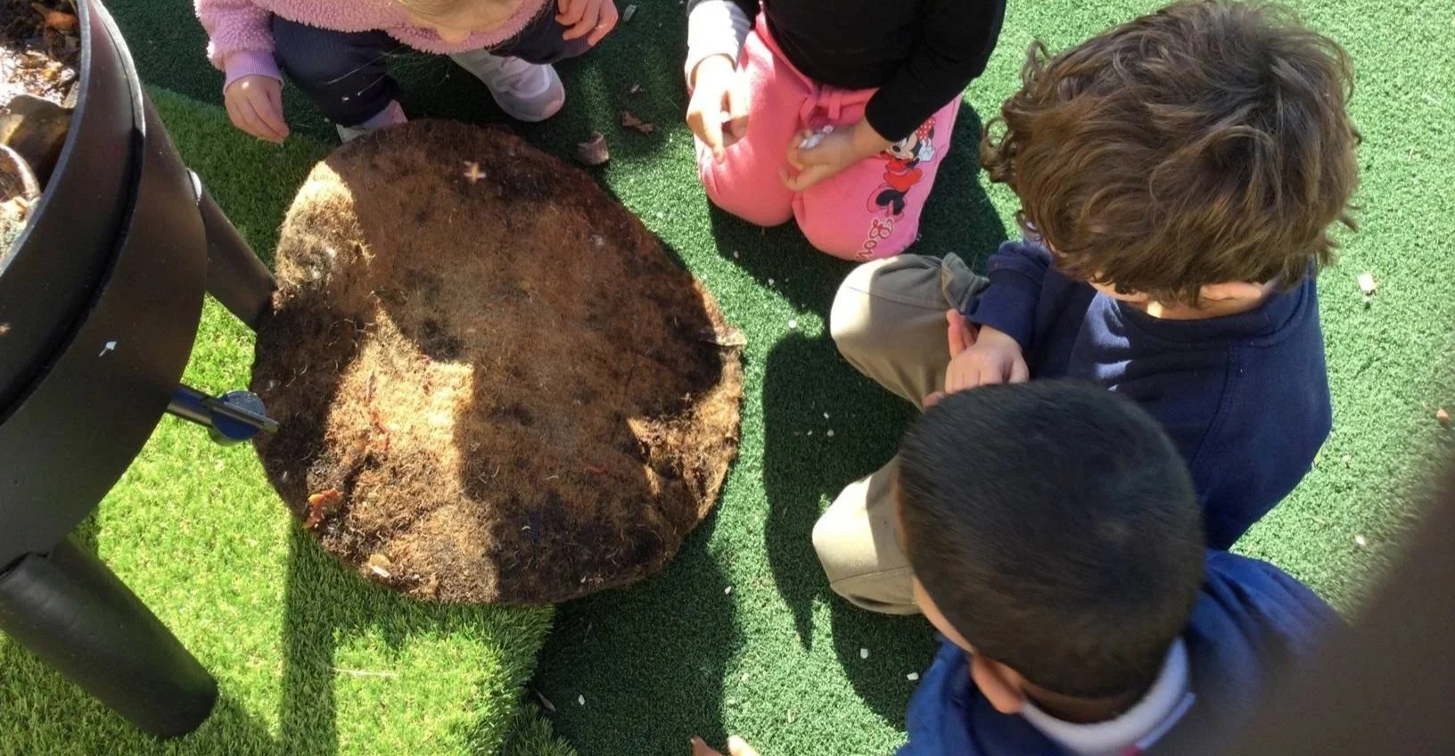 Children sitting outdoors on artificial grass, examining a large, round, felt object, possibly a tree root, part of a compost system, or buried item, near a black metal structure, possibly a compost bin.