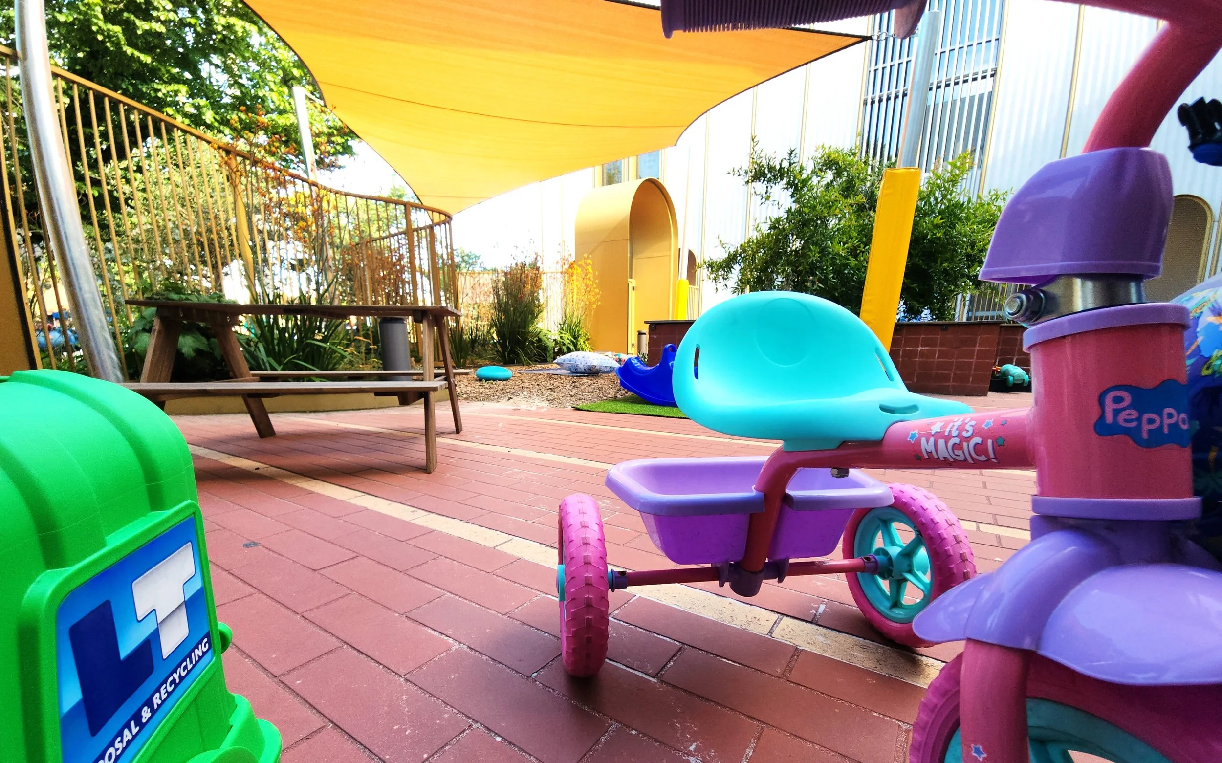 St Mary's Children Centre Kensington Victoria - outdoor play area featuring a pink tricycle, a blue rubber duck, a green recycling bin, a wooden picnic bench, and a yellow shade sail, surrounded by greenery and a modern building with glass panels.