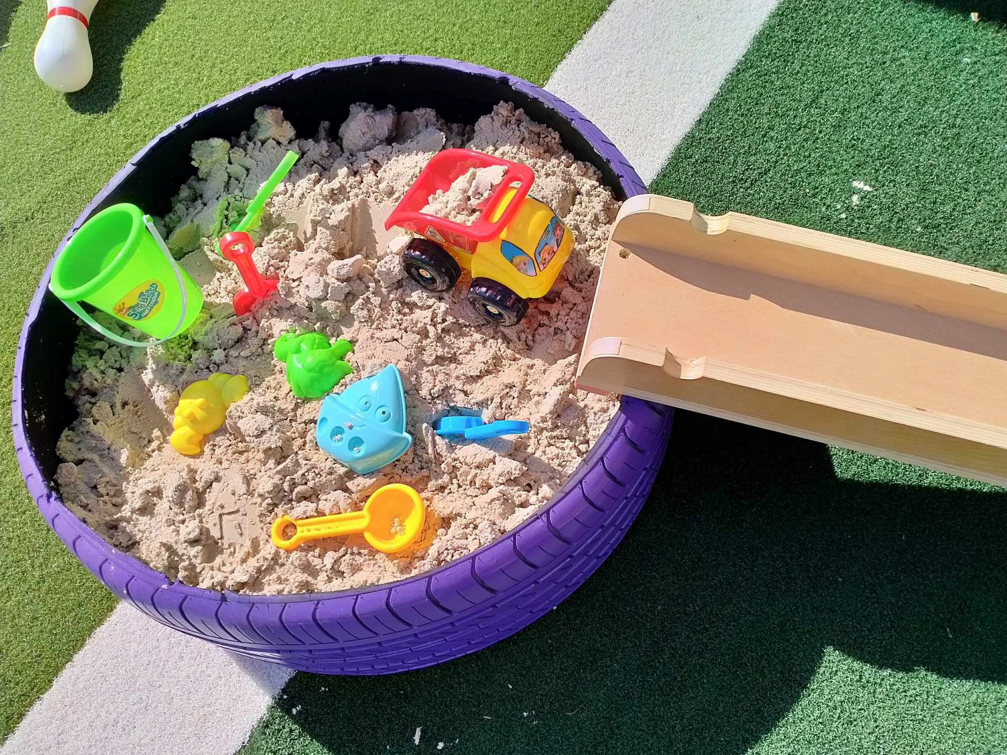 Children's recycled car tyre filled with sand, containing colorful play resources including a green bucket, a blue fish, a green shovel, a red truck, and more. The sand tyre is on a surface with green, gray, and black artificial turf.
