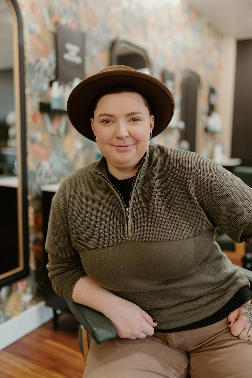 A person with short dark hair, wearing a brown hat and a green zip-up sweater, sitting in a salon or barbershop and smiling at the camera.
