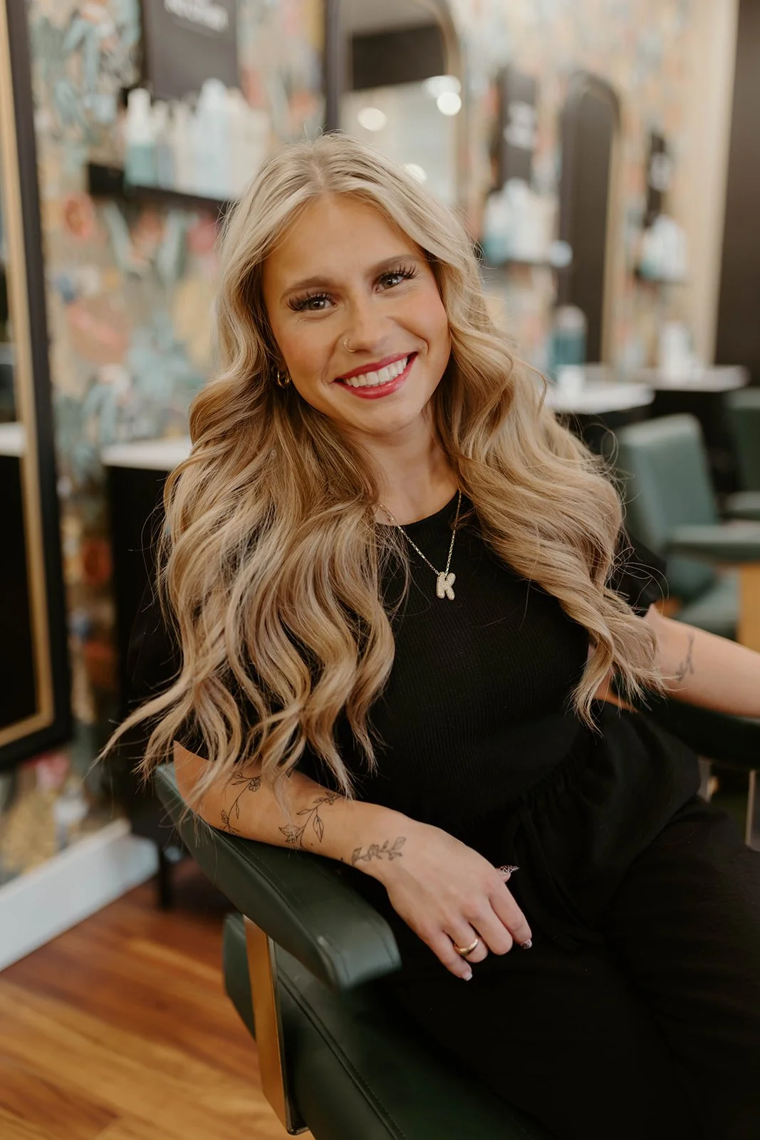 A smiling woman with long blonde curly hair sitting in a salon chair, wearing a black outfit with a necklace with a letter 'K' pendant.