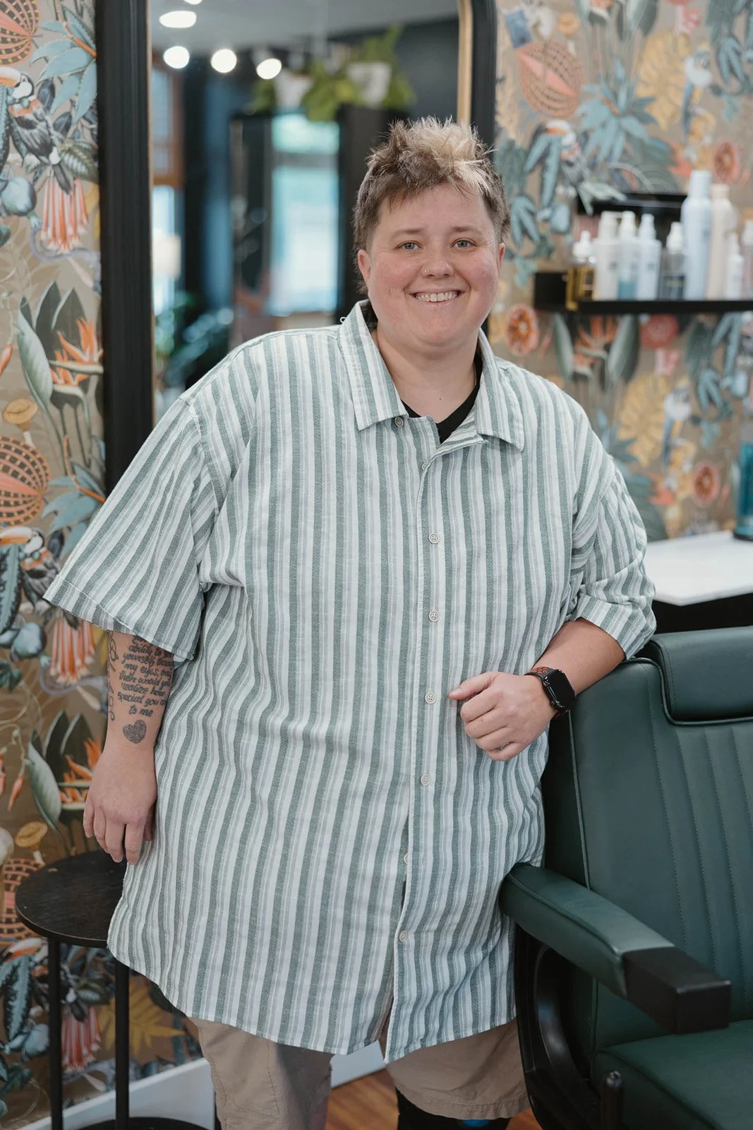 A smiling person with short dark hair styled upward, wearing a black jacket, sitting in a hair salon with floral wallpaper, mirrors, and other people in the background.